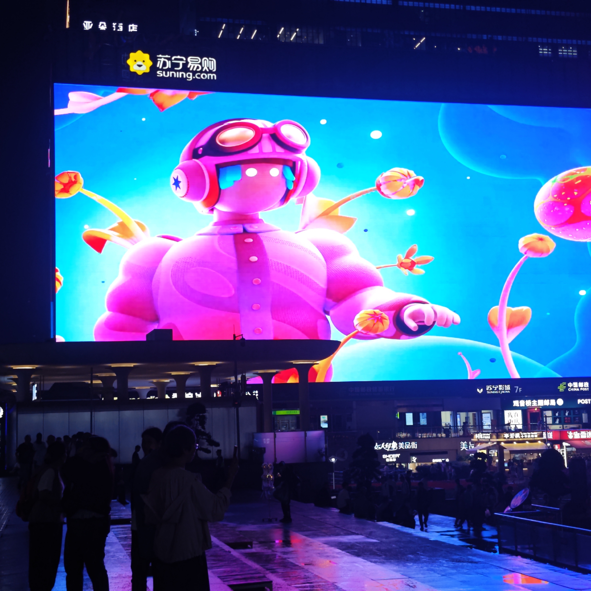 The image shows a nighttime scene in what appears to be a commercial area of Liangjiang, China. In the foreground, there are groups of people, partially obscured by shadows and possibly moving across the scene. Their actions and interactions are indistinct due to the image's focus. The ground is wet, reflecting the artificial lighting, creating a purple and blue hue on the walkway.
Dominating the background is a large LED screen displaying a colorful advertisement for "suning.com," a Chinese e-commerce company. The advertisement features a cartoonish character with goggles and protruding plants against a blue backdrop. Above the screen, partially visible signage includes characters that read "亚朵店" (Yaduo Hotel) and "苏宁易购" (Suning e-commerce).
Further back, shops and businesses line the street, with various Chinese characters indicating the names of the establishments. A sign for "中国邮政" (China Post) is also visible. The overall environment is bustling with pedestrian activity, illuminated by various sources of light that reflect off the wet surfaces, contributing to the scene's vibrant color palette.