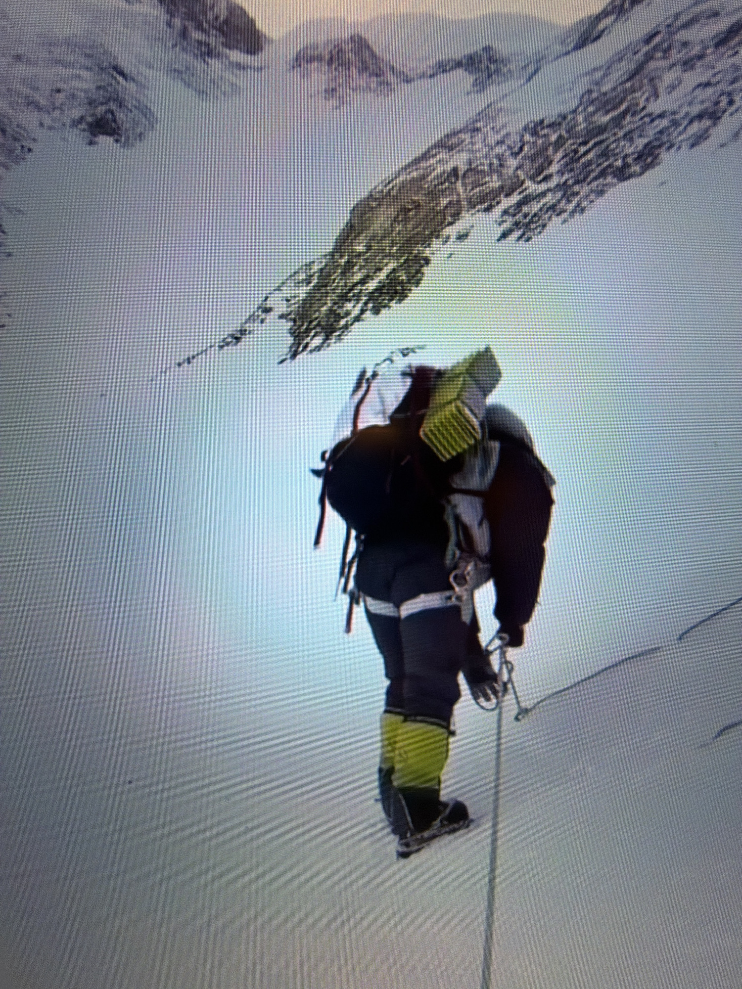 A person clad in winter mountaineering gear, including a black jacket, grey pants, and bright yellow boots, is ascending a snow-covered slope. The individual carries a large backpack with a yellow sleeping pad strapped to the top. A climbing rope is visible, held by the person and extending downwards. The background features a stark, mountainous landscape with snow and rock formations, suggesting a high-altitude, remote environment. There is no indication or discernible detail within the image that connects it to the city of Kafr Yasif, Israel.