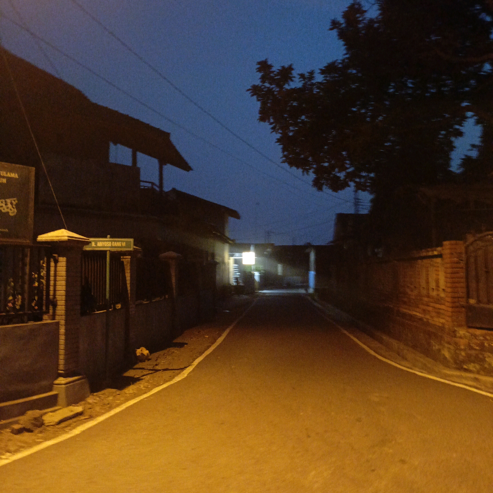 This is a nighttime photograph of a street in Salatiga, Indonesia, likely taken during twilight or early evening hours, as indicated by the deep blue sky and the presence of artificial lighting. The street is paved and has a yellow line marking its edge. On the left side of the street, there is a building with a dark roof and an overhanging structure. A fence runs along the front of the building, and a signpost with a yellow cap is visible. Another sign, illuminated by green light, reads "JL. ABIYOSO GANG VI". Further down the street, there appears to be a bright light emanating from a building on the left, illuminating the road ahead. The right side of the street features a brick wall. Silhouetted trees are visible against the darkening sky, and power lines crisscross above. The overall scene depicts a quiet, residential street.