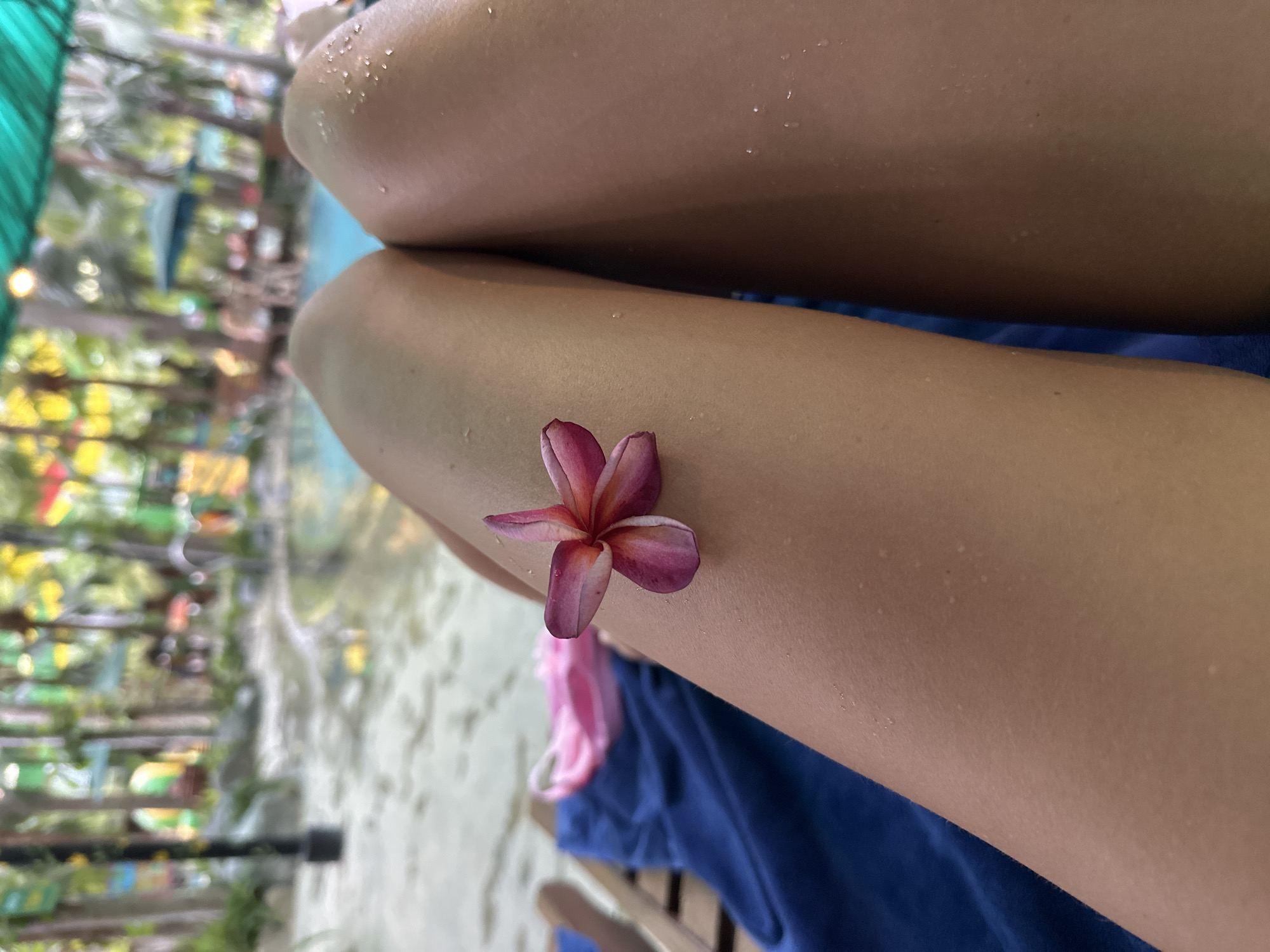 The image shows a close-up of a person's legs, likely a woman, with a pink flower resting on their thigh. The legs appear to be wet, suggesting they might be near water. A blue towel or piece of fabric is draped nearby. In the blurred background, there's a suggestion of an outdoor setting with vegetation and structures that may indicate a resort or recreational area. The location, Nam Nha Trang Ward, Vietnam, suggests a tropical or coastal environment, consistent with the image's ambiance. Specific details, such as age, clothing, or activity, are difficult to discern due to the image's focus and limited scope.