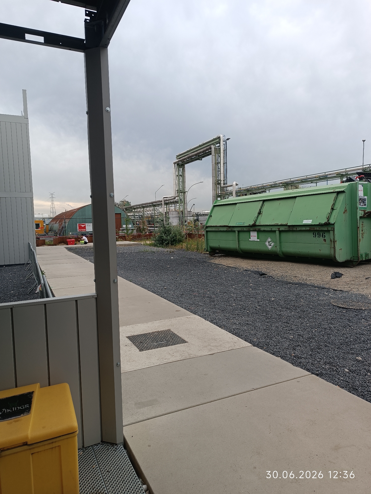 The image shows an outdoor industrial scene with a focus on a large green dumpster in the foreground, to the right of a concrete path. The dumpster has the number "996" painted on its side, as well as several hazard symbols and company logos. The path leads away from the viewer and curves to the right, bordered by dark grey gravel. To the left of the path is a grey modular building with vertical siding. Beyond the dumpster, industrial infrastructure is visible, including a complex network of pipes and elevated walkways, suggesting a factory or chemical plant. A rusted, barrel-shaped building with a sign reading "ALTIRA" is visible further in the background, as is a power pylon. The sky is overcast, indicating cloudy weather. In the far background, a person can be seen in the distance near the "ALTIRA" building. A yellow bin with the word "VIKINGR" is partially visible on the left side of the image. The timestamp "30.06.2026 12:36" is displayed in the bottom right corner of the image.