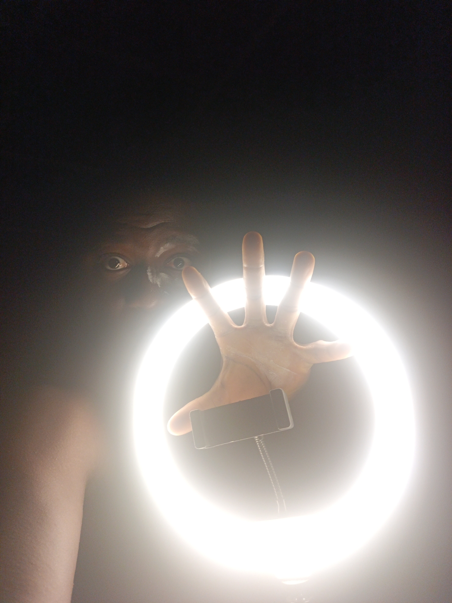 The image captures a person in a dark, undifferentiated indoor setting, illuminated dramatically by a large, bright ring light. The subject's face is partially visible on the left side of the frame, with wide, intense eyes peering from the darkness. There appears to be some dark substance, possibly paint or makeup, around their nose and eyes, enhancing a dramatic or theatrical appearance.
A bare shoulder and upper arm are visible on the far left. The person's right hand is outstretched into the frame, positioned in front of the ring light, with fingers slightly splayed and illuminated from behind, creating a silhouette effect around the edges of the hand.
Within the bright circle of the ring light, a dark smartphone holder or clamp is visible, positioned towards the bottom center of the light. A cable, possibly for power or audio, can be seen connected to this holder or the light itself. The extreme brightness of the ring light creates a stark contrast with the surrounding darkness, making most of the background indistinguishable. The direct gaze towards the camera and the setup with the ring light and phone holder suggest the person is either taking a selfie, participating in a video call, or recording content. No visible text is present.