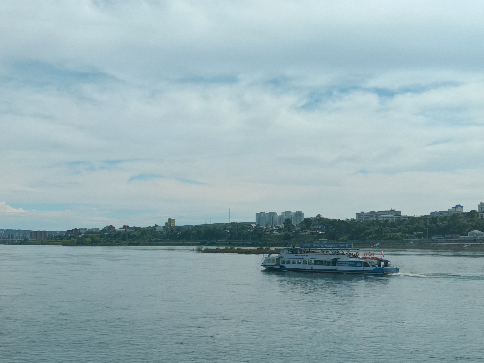 A wide, calm river flows in the foreground, its surface rippled by a gentle breeze. On the right side of the river, a white passenger boat with blue trim is moving to the right, leaving a white wake. The boat is populated with many passengers visible on its upper deck. The far bank of the river features a cityscape with a dense arrangement of buildings, varying in height and architectural style, nestled amongst lush green trees. Some taller, modern apartment buildings stand out, while lower-rise structures and industrial elements are also visible. The sky above is overcast with light blue and white clouds, indicating a daytime scene. The overall impression is a view of a city on a river during fair weather. The provided text on the boat, "ТЕплоход МОСКВА-4", indicates it is a type of motor ship commonly used for river cruises. Based on similar views, this scene likely depicts the Angara River in Irkutsk, Russia, with the city skyline visible on the left bank.