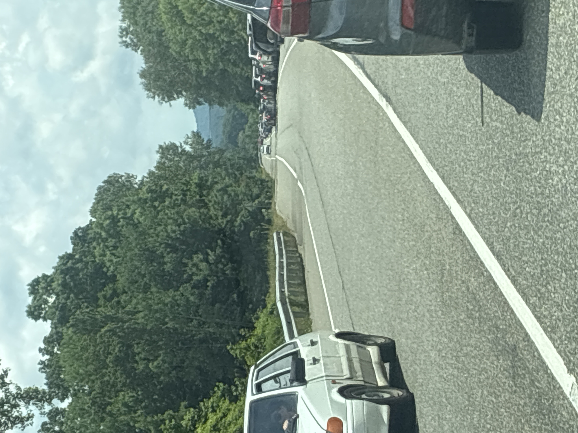A white car is in the foreground on the left side of the image, partially obscuring the view of a traffic jam. The car has its passenger door open and a person's arm is visible inside. The road is paved and has white lane markings. The traffic jam consists of numerous cars of various colors, stretching into the distance. The road is lined with dense green trees on both sides, and hills can be seen in the background. The sky is overcast with clouds. The image appears to be taken from a moving vehicle. The traffic jam is on a two-lane road, likely in a rural or semi-rural setting outside of Tekos, Russia.