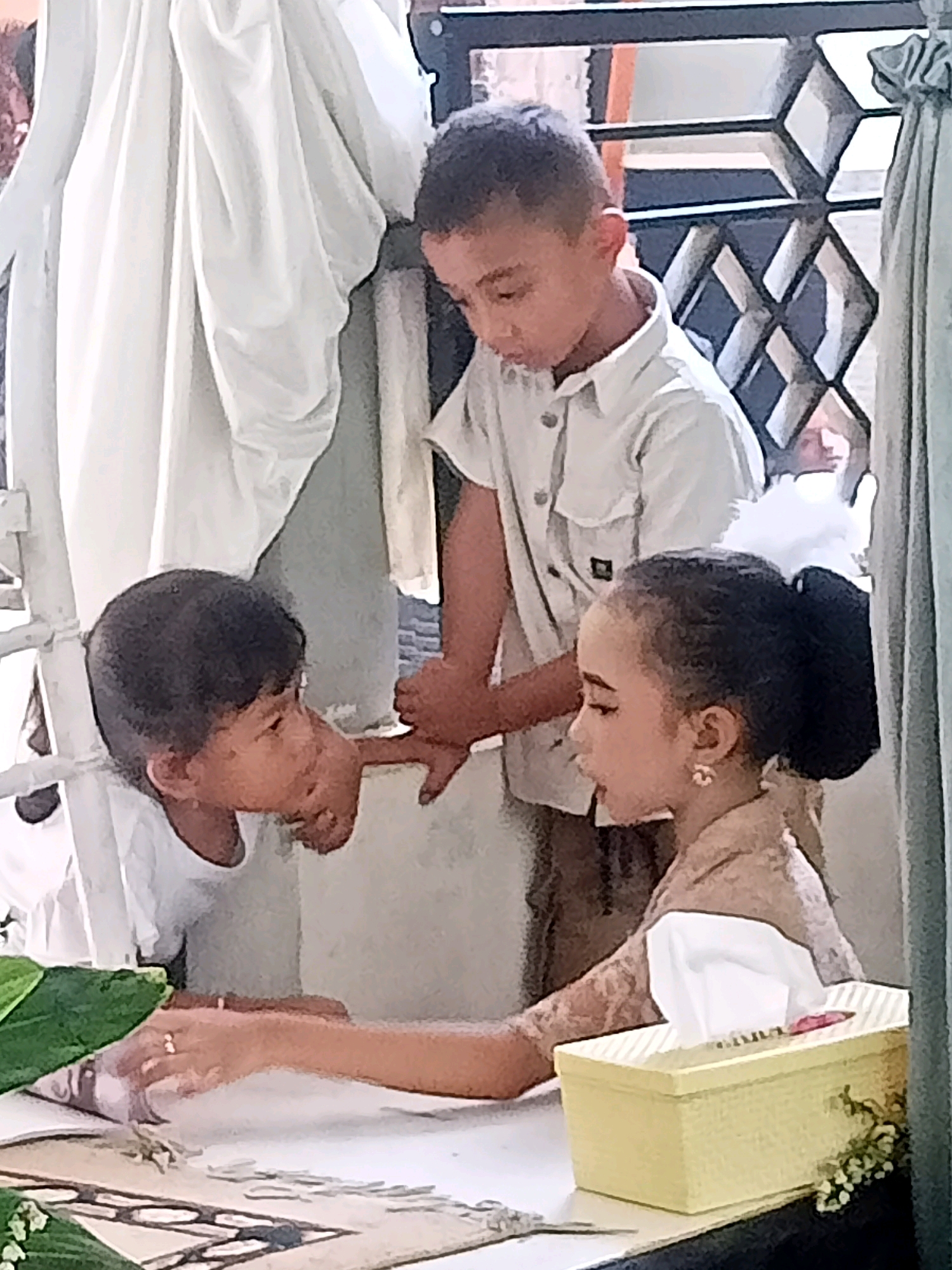 In Badamita, Indonesia, three children are gathered around a table. A young boy, wearing a white collared shirt, leans in, with his right hand extended as if to touch something on the table. Facing him is a young girl with her hair pulled up in a bun and wearing a light-colored top with lace detailing. She is looking down at the table. Standing behind the boy is another boy, also in a white collared shirt, who appears to be observing the interaction. The table is covered with a white cloth, and a yellow tissue box with a floral decoration sits to the right. To the left of the table, a green leafy plant is partially visible. The background features a white curtain on the left and a patterned metal screen and a green curtain on the right.