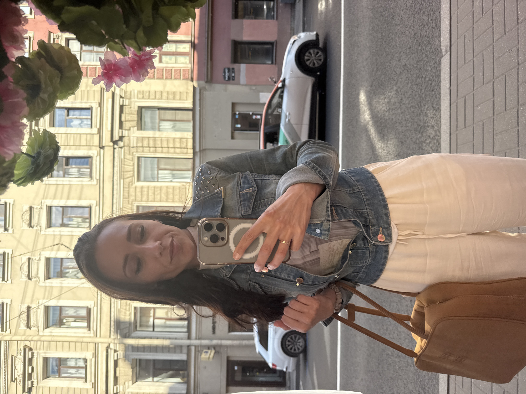 A woman with long, dark hair takes a selfie in Saint Petersburg, Russia. She is wearing a denim jacket with silver studs on the shoulders and a light-colored, sheer top. Her lower half is clad in loose-fitting, cream-colored trousers. She carries a tan leather shoulder bag. In the background, a white SUV is parked and a silver car with green and orange accents is driving by on the street. The architecture in the background is typical of Saint Petersburg, with ornate building facades and large windows. Pink and green artificial flowers hang in the foreground, partially obscuring the view. The scene is set on a city sidewalk, with a paved street and sidewalk visible.