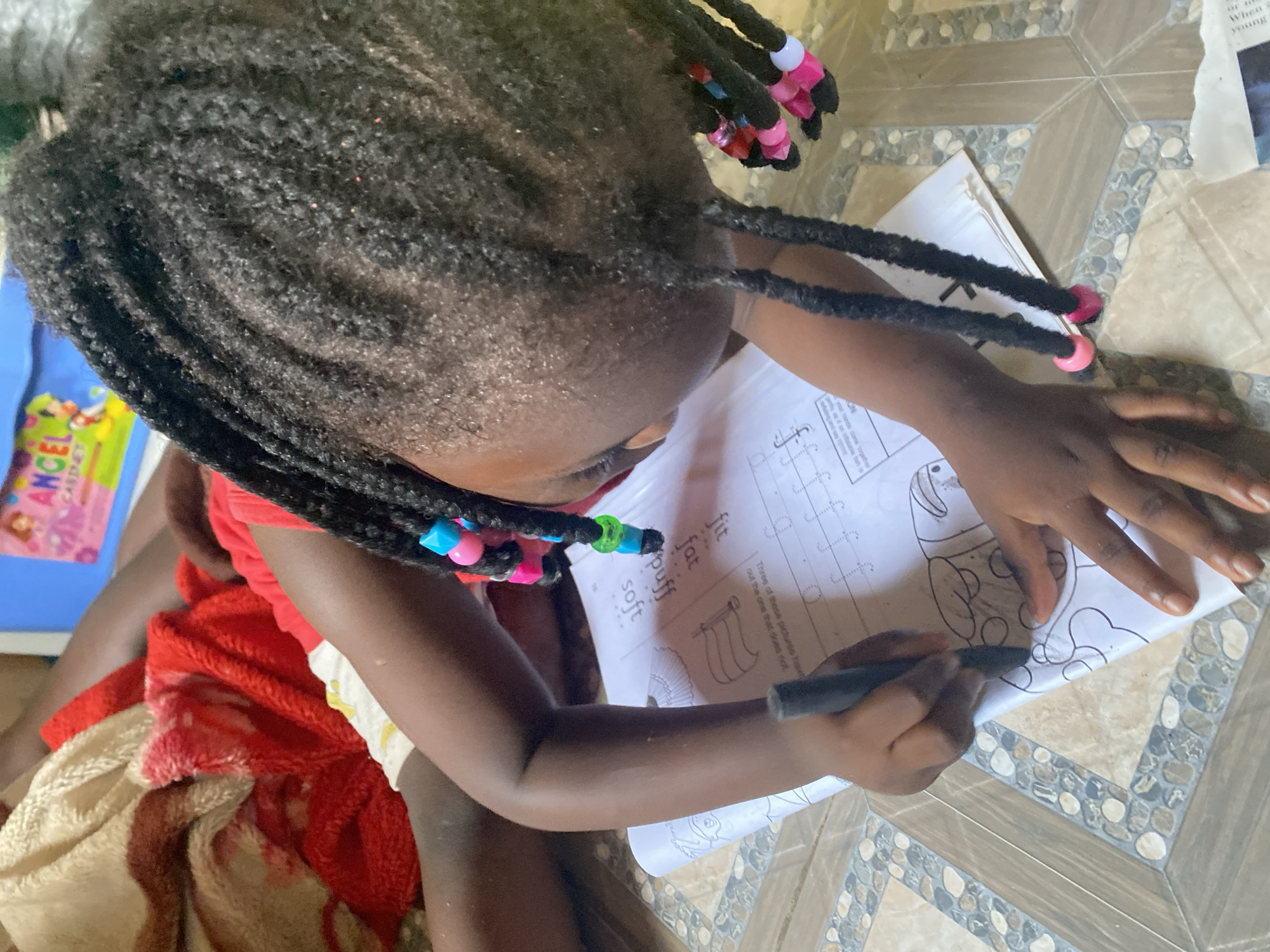 A child is sitting on a tiled floor, coloring in a coloring book. The child has dark hair styled in braids with colorful beads. The child is wearing a red shirt and appears to be sitting on a blanket. The coloring book has words on the page: "fit," "fat," "puff," and "soft." Next to the child, there is a "CABINET ANGEL" book. The location is Rido, Nigeria.