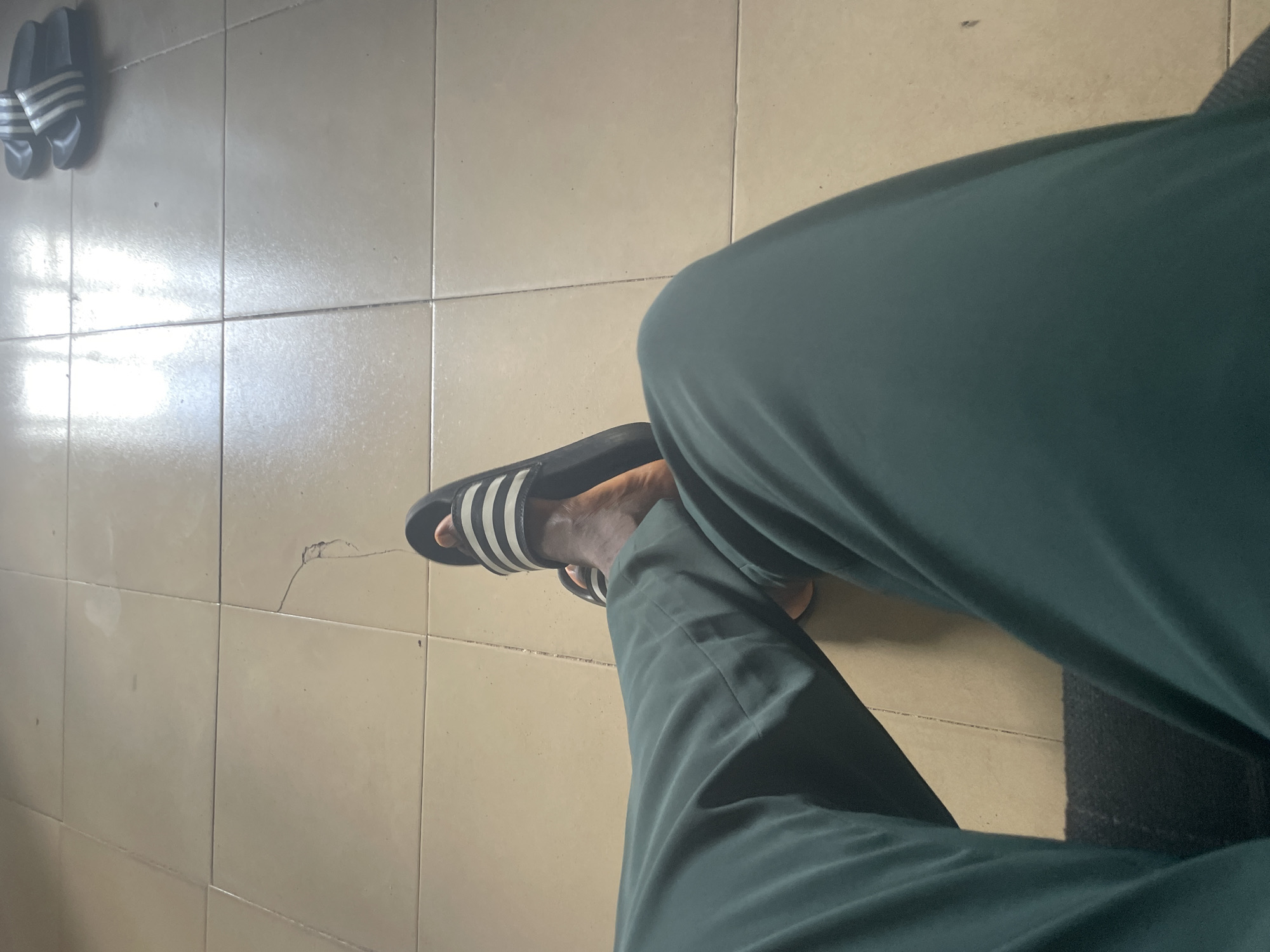 A person's legs are visible, wearing dark green pants and black and white striped slides, resting on a tiled floor in what appears to be a residential setting in Jalingo, Nigeria. The tiles are light beige and show some wear, with a noticeable crack in one of the tiles. The lighting suggests an indoor environment, possibly a living room or hallway, with light reflecting off the tiles from a window or overhead light source. There are no other people or significant objects in the frame.