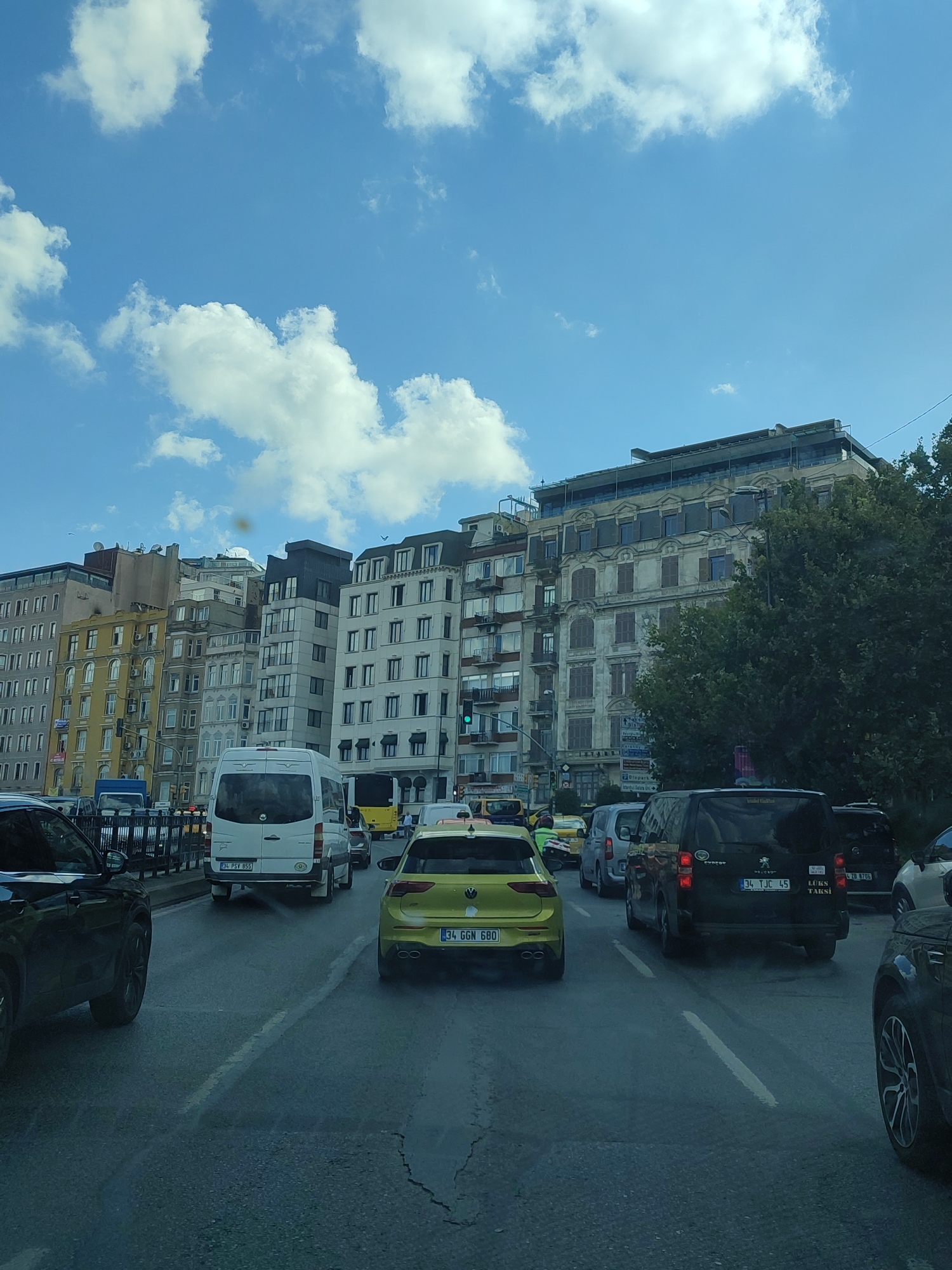 The image shows a street scene in Beyoğlu, Turkey, with heavy traffic under a bright blue sky with white clouds. Several cars, vans, and a bus are visible on the road, indicating a busy urban environment. The buildings lining the street are multi-story structures with varying architectural styles, some appearing older and others more modern, suggesting a mix of historical and contemporary development. Trees are also present, adding a touch of greenery to the cityscape. The overall impression is one of a typical day in a bustling Turkish city, with vehicles and pedestrians navigating the urban landscape.