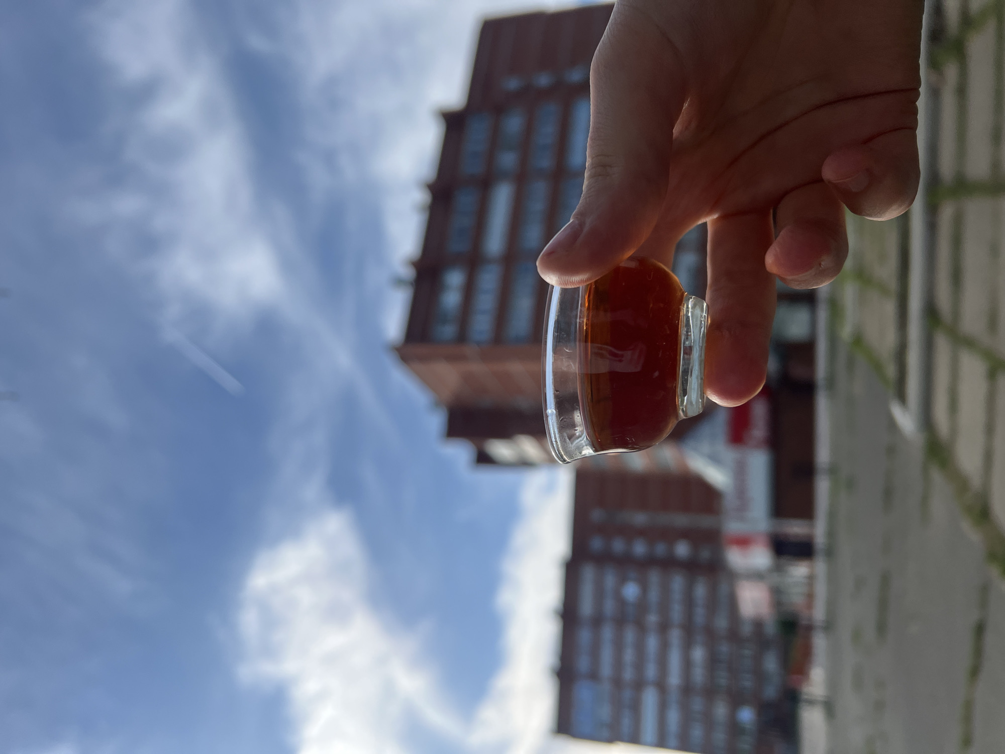 A hand holds a small glass filled with a brown liquid in front of an out-of-focus cityscape. The background features a cloudy blue sky and two tall, brown buildings with repetitive window patterns. Part of a gray, paved surface is visible at the right of the frame. The photo appears to be taken outdoors during daylight. The location is in the city of Chaikovsky, Russia.