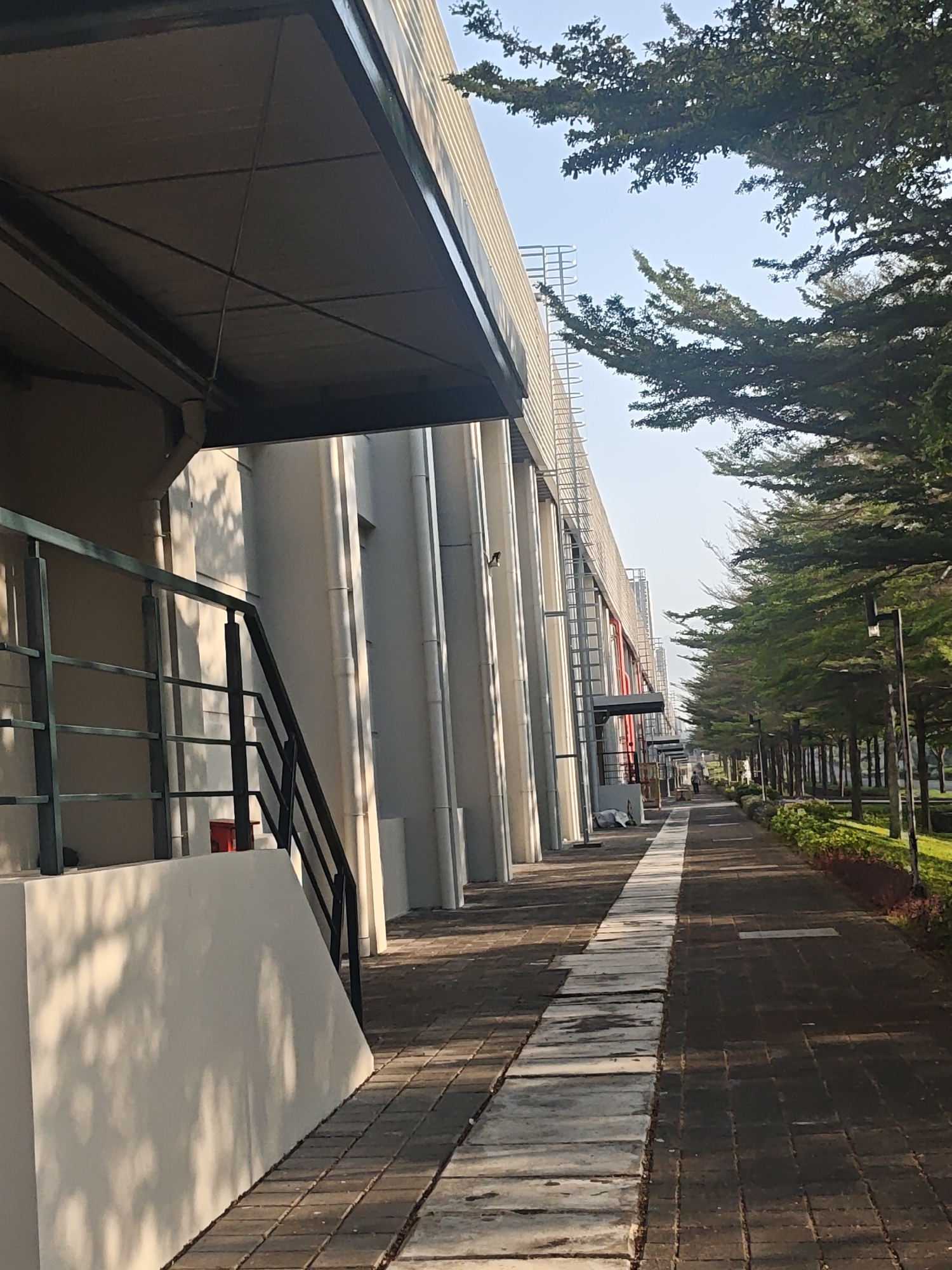 This is a vertical, eye-level shot of a paved walkway beside a modern building and a line of trees in Bae, Indonesia. The building, on the left, features a metal overhang supported by white columns and a black metal railing alongside a staircase. The walkway is paved with dark rectangular bricks, with a strip of lighter concrete slabs running down the center. On the right, a row of evenly spaced trees with lush green foliage lines the walkway, with manicured bushes and red mulch at their base. A black lamppost is visible among the trees. The sky is clear and blue, with the sun casting shadows from the trees and building onto the walkway and the building's facade. In the distance, a single person is walking away from the camera on the walkway.