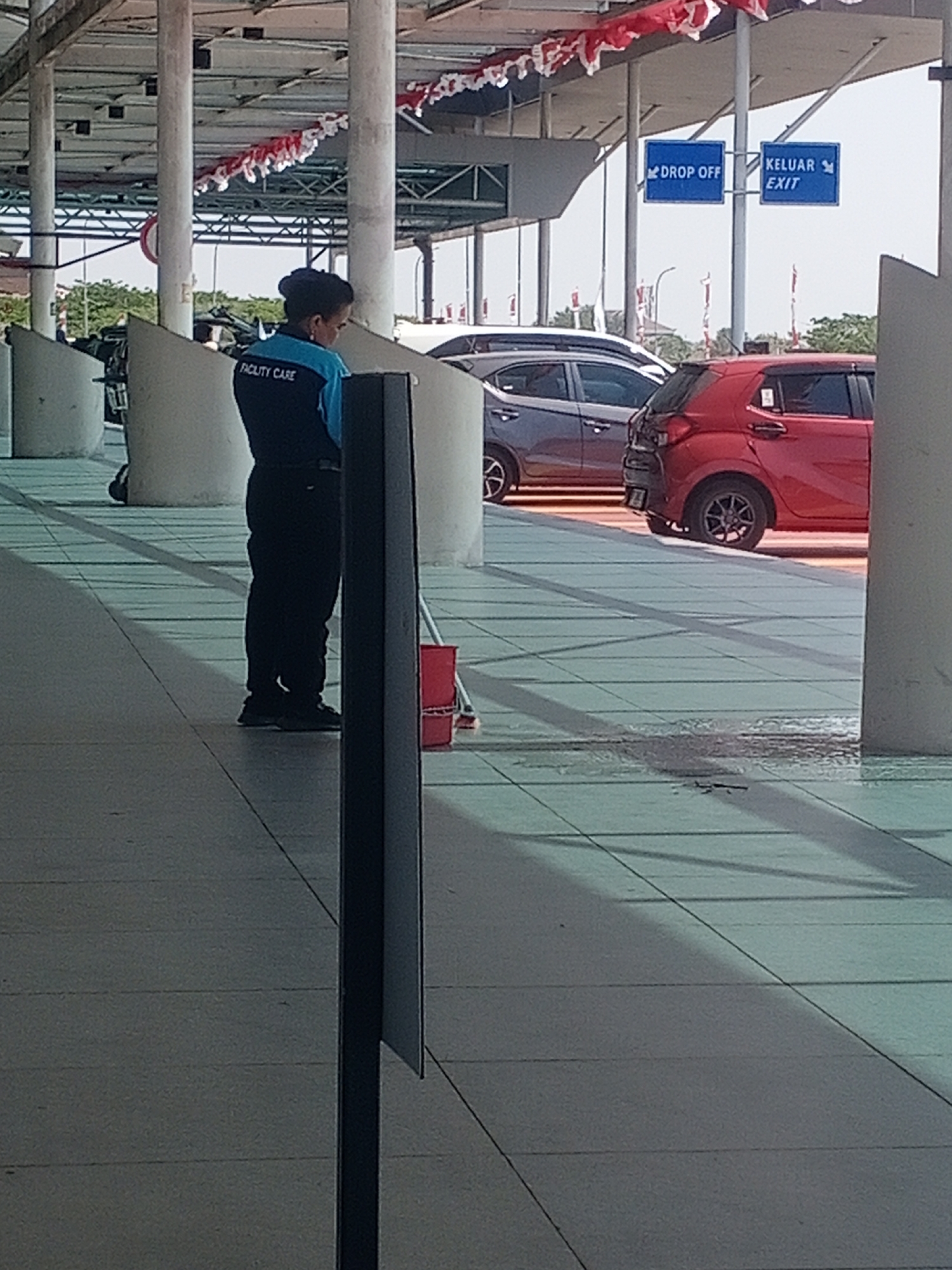 A person wearing a blue shirt with "FACILITY CARE" printed on the back and black pants is standing in what appears to be an outdoor covered area with a tiled floor. The person is facing away from the camera, and their stance suggests they might be cleaning or attending to something on the ground. A red bucket and a mop are visible near their feet, indicating cleaning activity. In the background, there are signs for "DROP OFF" and "KELUAR EXIT" on blue rectangular signs, suggesting a transportation hub or public facility. Several cars are parked or driving in the background, including a red car and a darker colored sedan. The environment is bright and appears to be daytime, with sunlight casting shadows on the tiled floor. Decorative red and white banners are strung across the ceiling structure. The location is identified as Baji Mangngai, Indonesia.