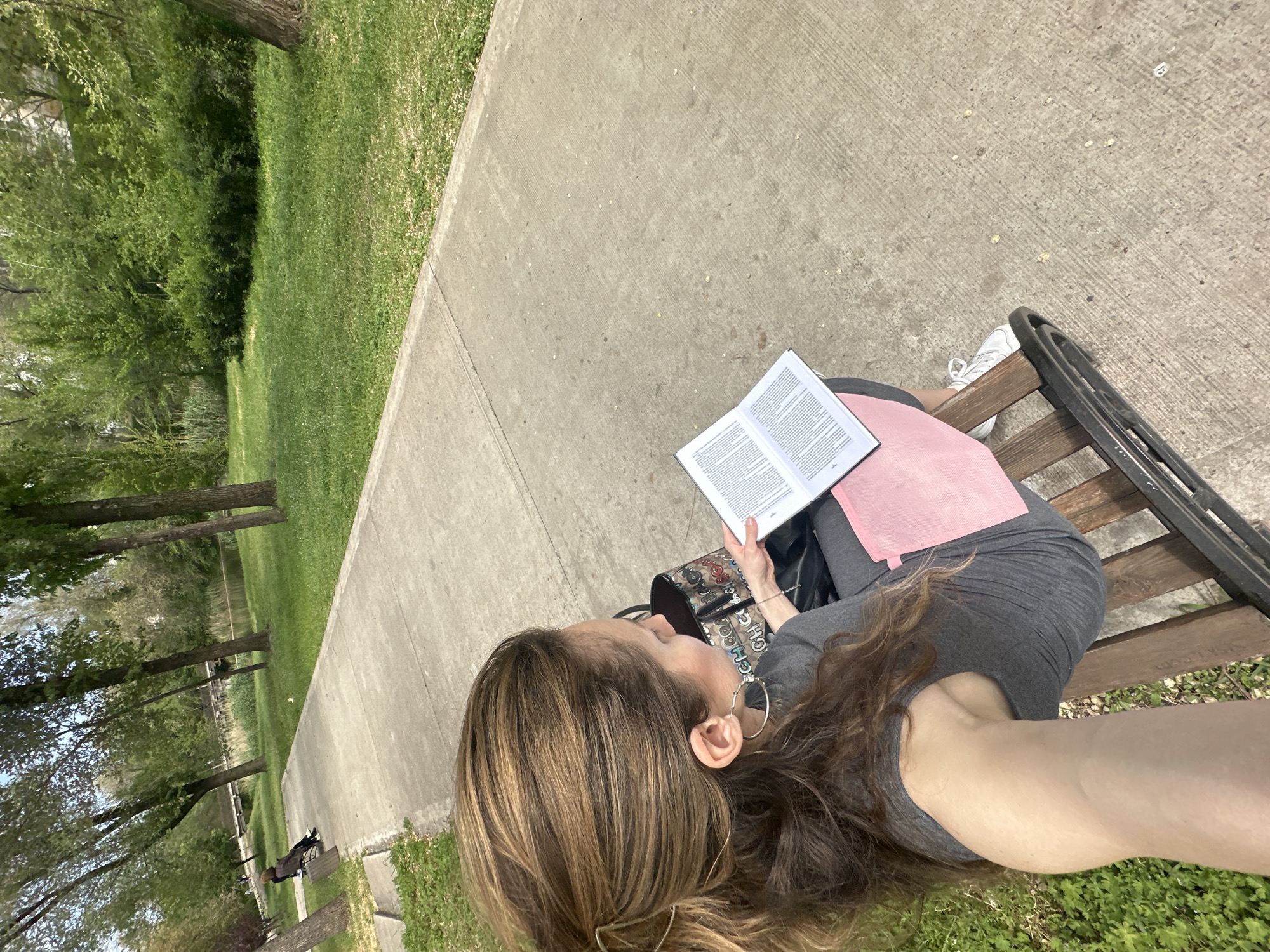 A woman is sitting on a park bench outdoors, reading a book. She is wearing gray clothing and has long, highlighted hair. She is holding the book open with both hands. A pink object rests on her lap. Her purse or bag is to her left. The park bench is situated next to a concrete path that runs parallel to a grassy area with trees. A lake can be seen through the trees in the background. The location appears to be a park or green space. The city of Kryvyi Rih, Ukraine cannot be positively identified from the visual information provided.