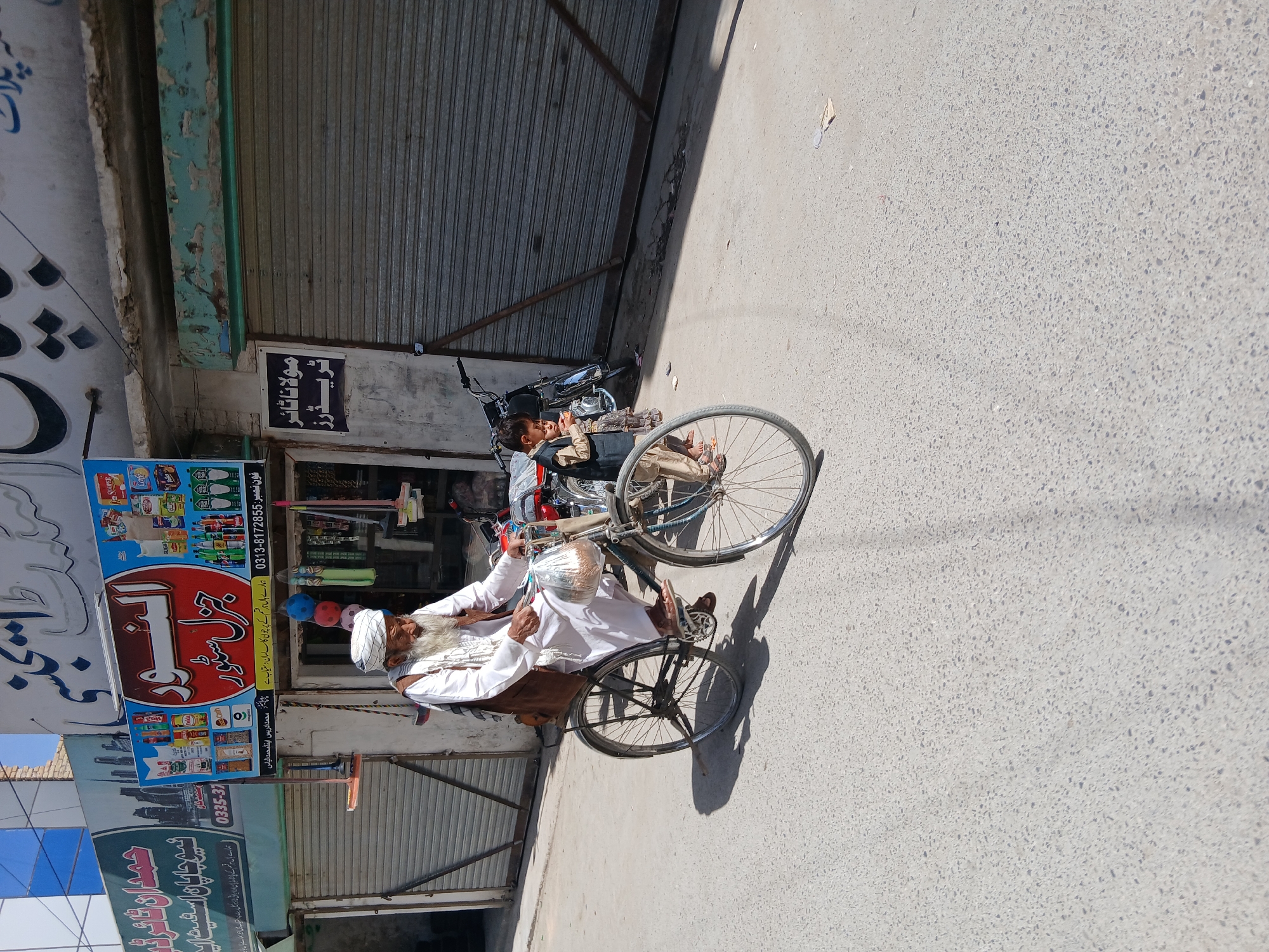 The image shows an elderly man, likely a local resident, sitting on a bicycle rickshaw in front of a store in Quetta City Tehsil, Pakistan. The man is wearing traditional attire, including a turban and long, flowing garments. A young boy is riding on the back of a parked motorcycle next to the rickshaw. The store behind them has signage written in Urdu or Arabic and appears to be a general store selling various items, including food and household goods. The overall environment is a street scene with concrete pavement and buildings on either side.