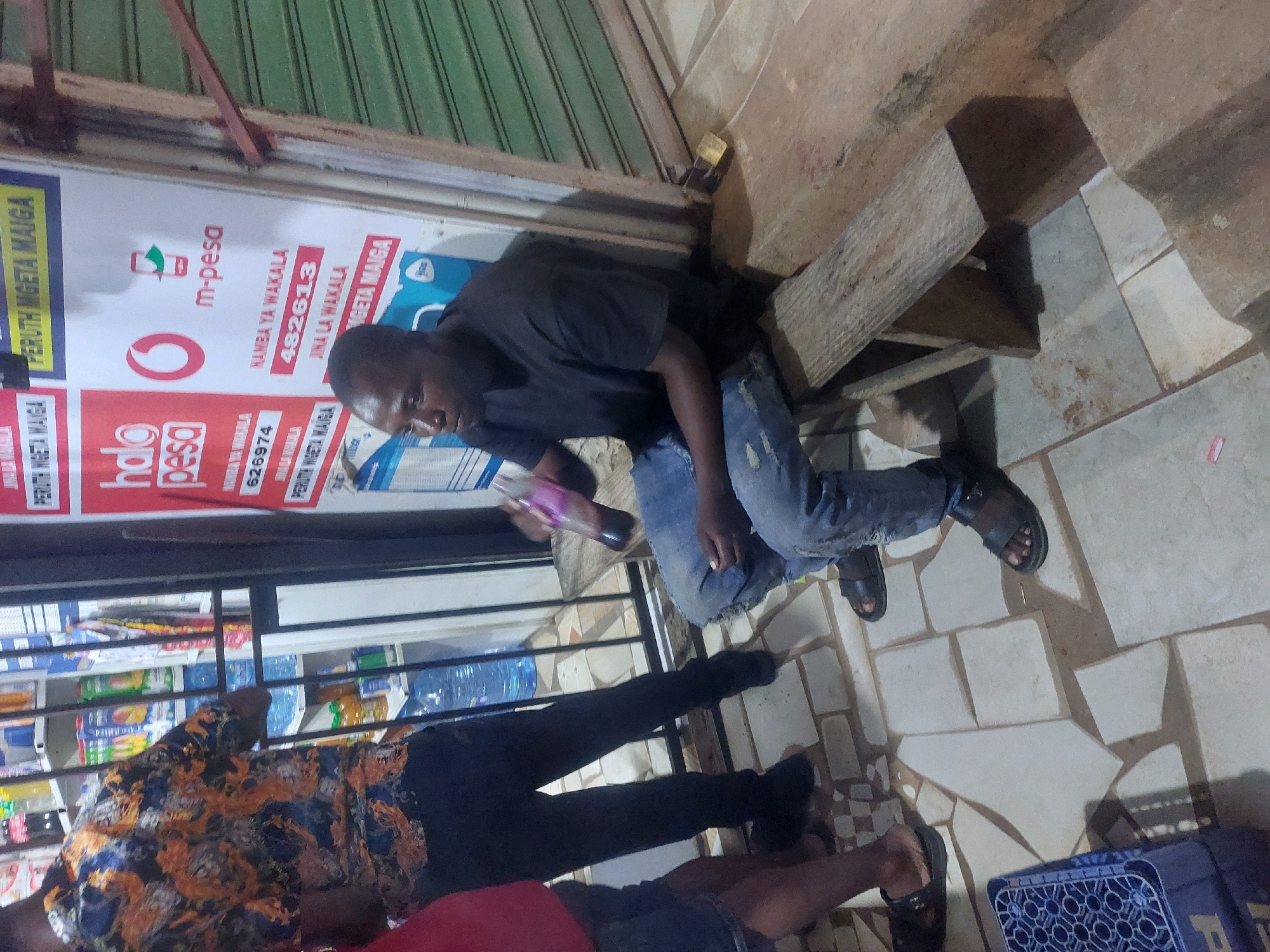 The image shows a man in a black shirt and jeans sitting on a makeshift wooden bench in front of a shop in Dar es-Salaam, Tanzania. The shop has signs for m-pesa and halo pesa. The m-pesa sign includes "NAMBA YA WAKALA 482613 JINA LA WAKALA MGETA MAIGA," indicating it is an m-pesa agency owned by someone named Mgeta Maiga, with the agency number 482613. Similarly, the halo pesa sign says "NAMBA YA WAKALA 626974 PERUTH INGETA MAIGA". He is holding a bottle. Other people are present, and a blue crate is on the ground. The floor is tiled. The shop appears to be closed, with the shutter down.