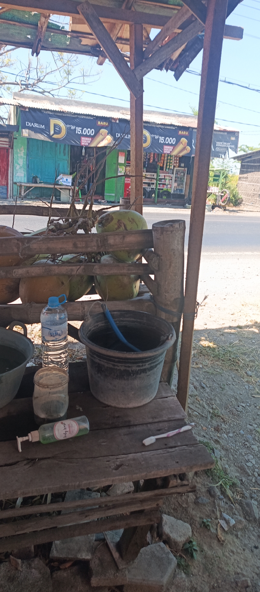 A roadside stand in Jatisari, Indonesia, is captured in a medium shot. Coconuts are piled on a wooden structure in the foreground. A bottle of water, a plastic jar, a bottle of green liquid, and a bucket with a blue hose are on a wooden plank. In the background, buildings with green doors and banners display advertisements. The scene is under a wooden roof, and the ground is covered with dirt and rocks.
