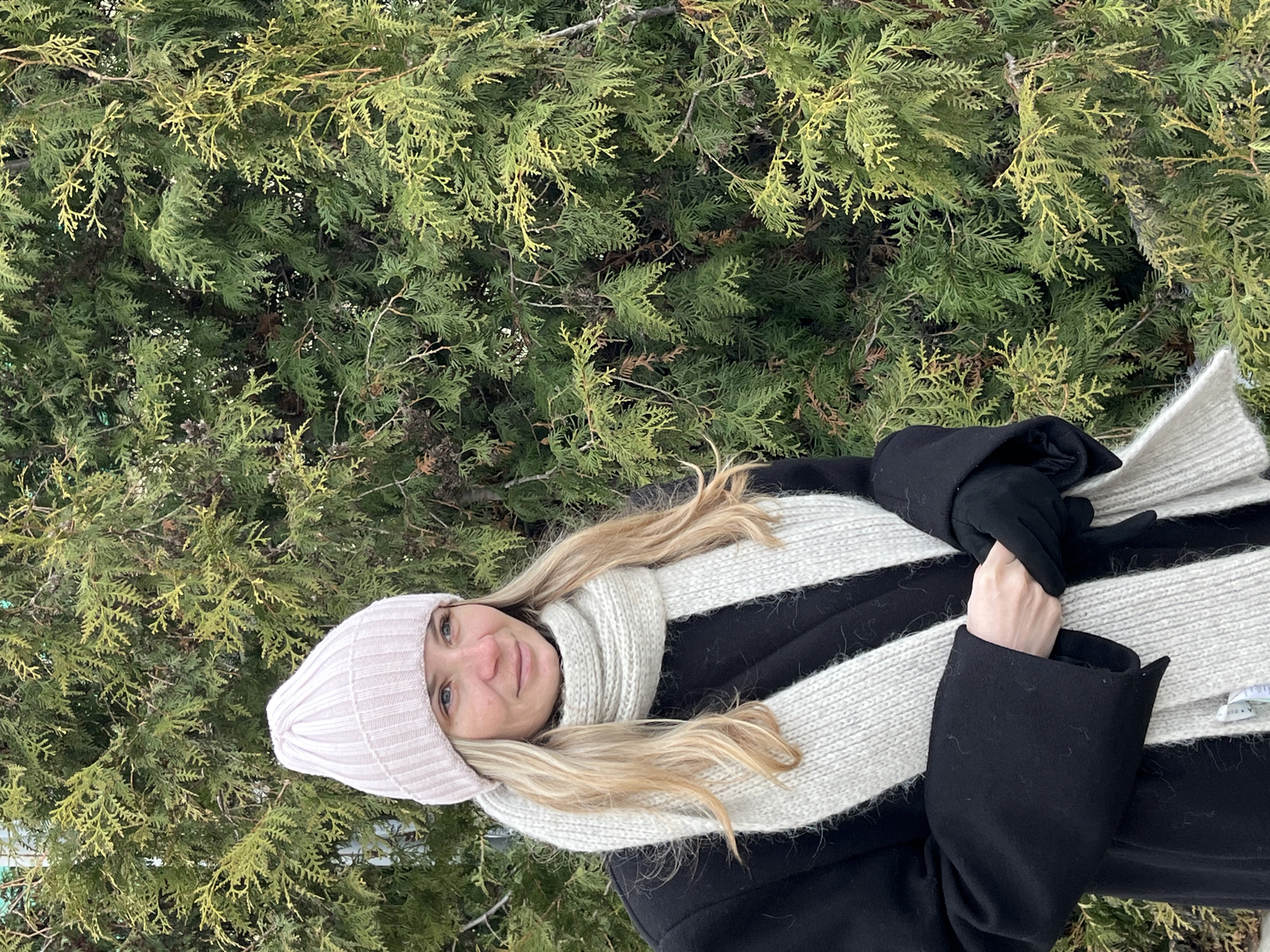A woman is posing for a photograph in front of a dense evergreen shrub or tree. She is wearing a pale pink knit hat, a white and black striped scarf, black gloves, and a black coat. She has blonde hair. The location appears to be outdoors. There is no definitive information in the image to confirm the location is in Chaikovsky, Russia.