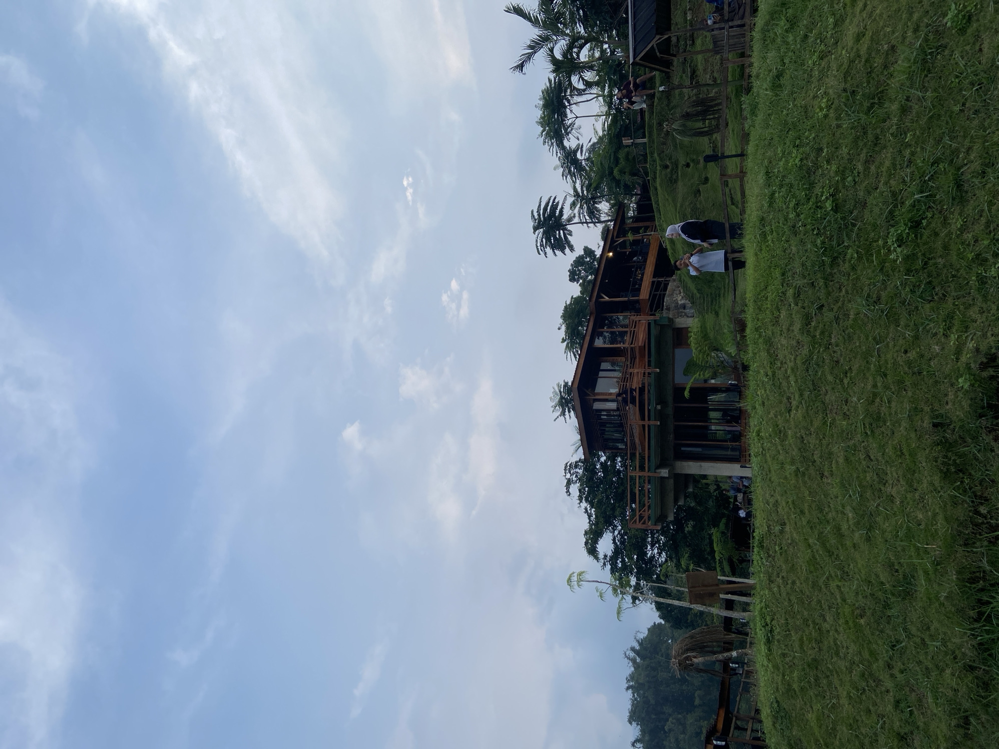The image depicts a daytime scene in Tamiajeng, Indonesia. A two-story wooden building with glass windows and a wooden balcony is situated on a grassy hill. A few individuals are standing on the balcony and in the surrounding grassy area. Tall palm trees are visible behind the building. The sky is light blue with scattered white clouds. Lush green vegetation covers the hill. A small, thatched-roof structure is partially visible in the lower left corner.