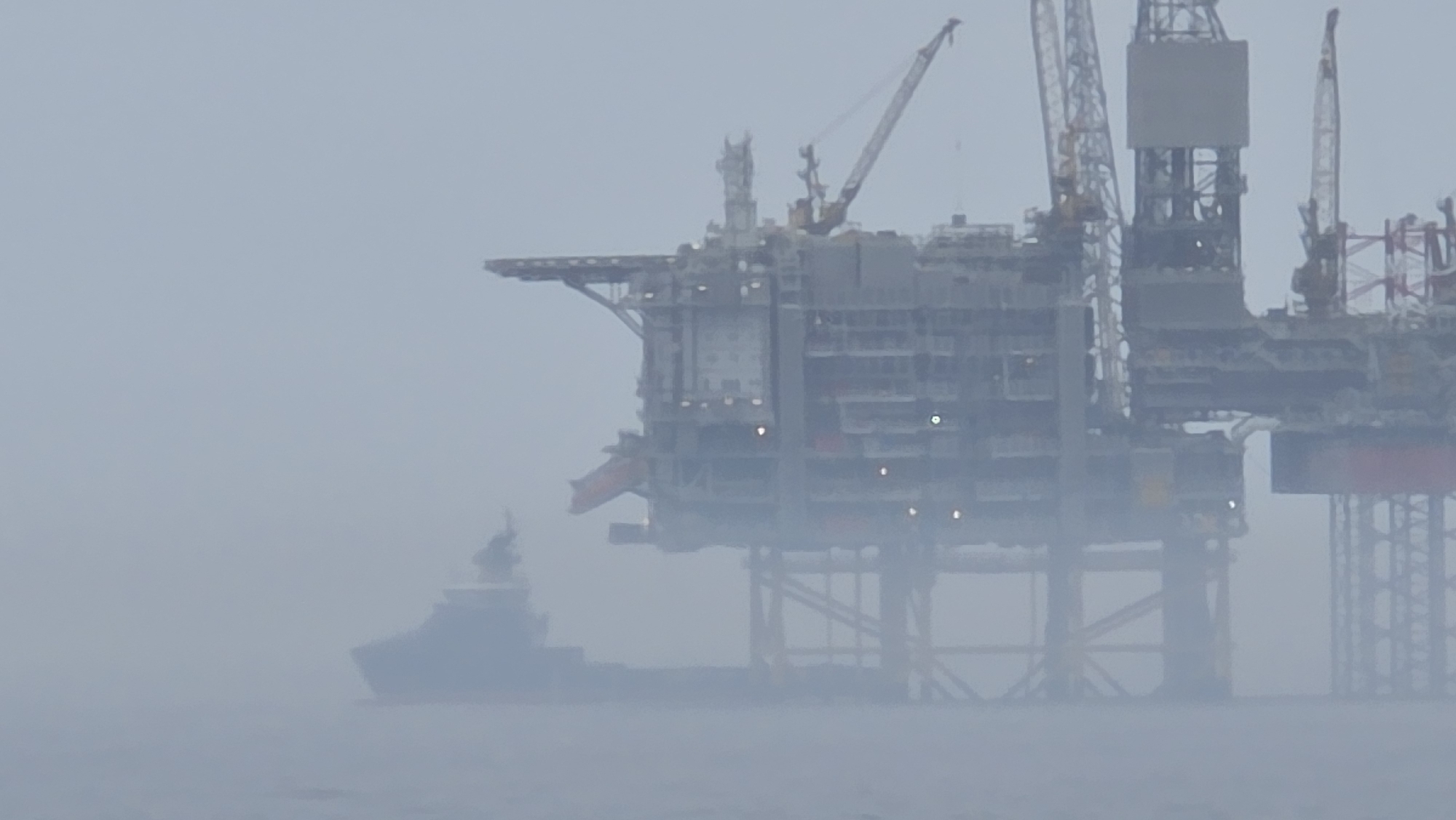The image depicts an oil platform and a ship in a foggy environment. The platform is a large, complex structure with multiple levels, cranes, and support beams. The ship is smaller in scale and located to the left of the platform. The overall scene is obscured by fog, reducing visibility. Location: Unknown, Unknown.