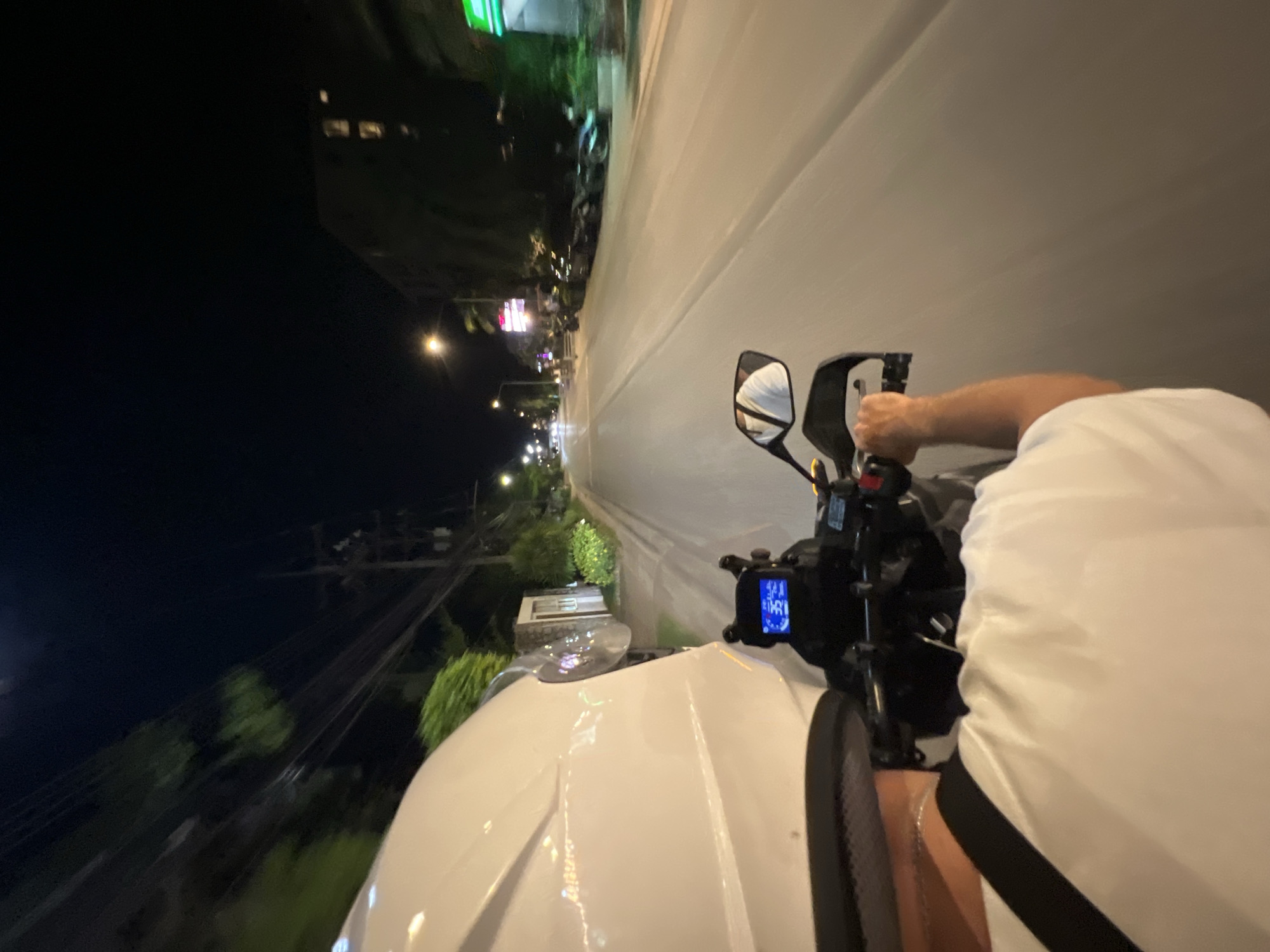 A tilted, nighttime shot from the first-person perspective of a person riding a motorcycle. The motorcycle is white, and the rider is wearing a white shirt with a black stripe. The view includes the dashboard, handlebars, and side mirror. The motorcycle is moving along a well-lit road, with city lights and buildings visible in the background. The location is in the city of Unknown, Unknown.