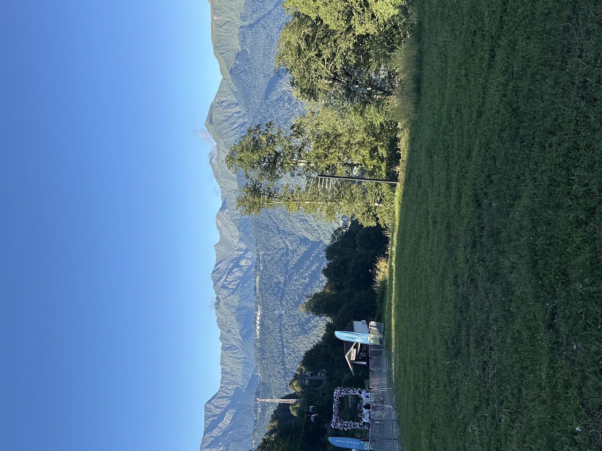 The image depicts a mountain landscape near Estosadok, Russia, under a clear blue sky. In the foreground, a grassy slope descends towards a fenced area with several people gathered. Behind the fence, there's a floral archway, a small building, and two blue flags. A ski lift with gondolas is visible in the mid-ground, ascending a treed hillside. The background is dominated by layered mountain ranges covered in dense green forest, with snow-capped peaks in the distance. The overall scene suggests a summer or early autumn day at a mountain resort.