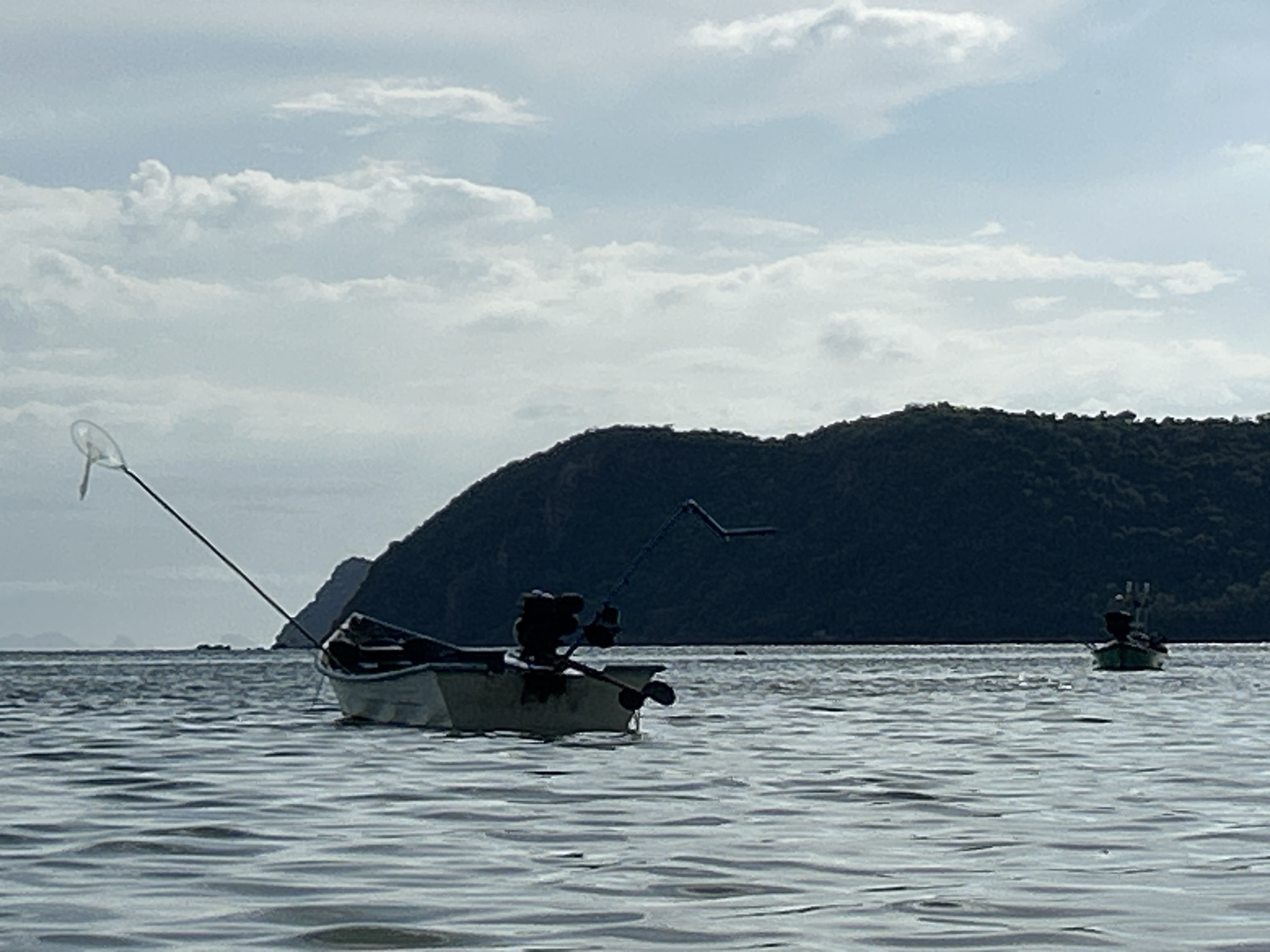 The image shows two small boats on the water near ตำบลเกาะพะงัน, Thailand. The first boat is white and has a motor, oars, and a net attached to a pole. The second boat is green. A mountain is visible in the background. The sky is cloudy.