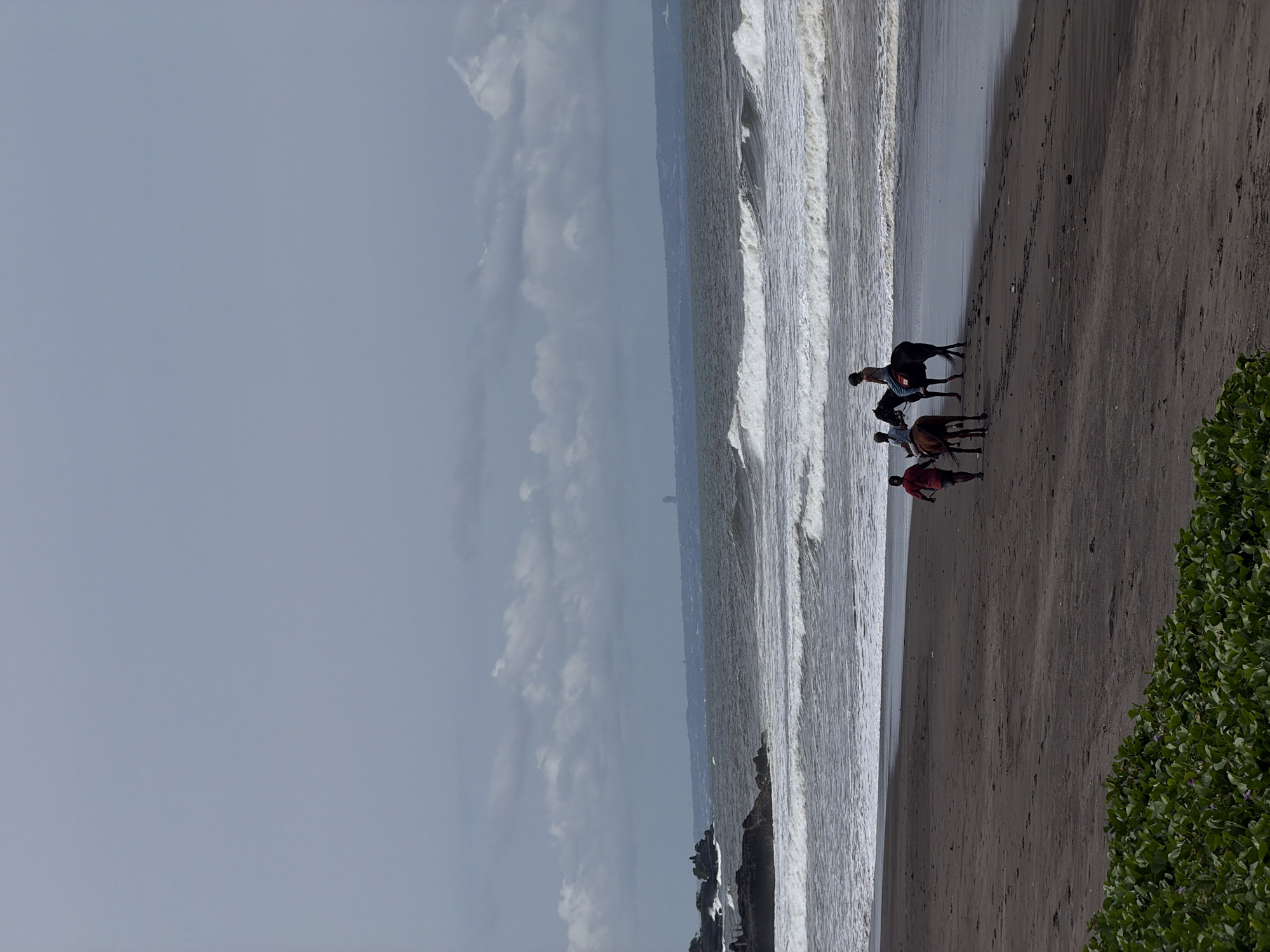 A dark sand beach in Belalang, Indonesia, is depicted under an overcast sky. Three individuals and two horses are positioned near the ocean's edge. One person in a red shirt walks on the sand, leading a dark brown horse. To their right, two other individuals, each wearing a light-colored top, dark pants, and a helmet, are mounted on separate dark brown horses, riding parallel to the shoreline. Moderate ocean waves are breaking onto the beach. Rocky formations are visible in the ocean on the far left. A distant coastal landmass, featuring structures consistent with human habitation and a distinct temple, is visible across the water. Green ground cover is present in the lower-left foreground.