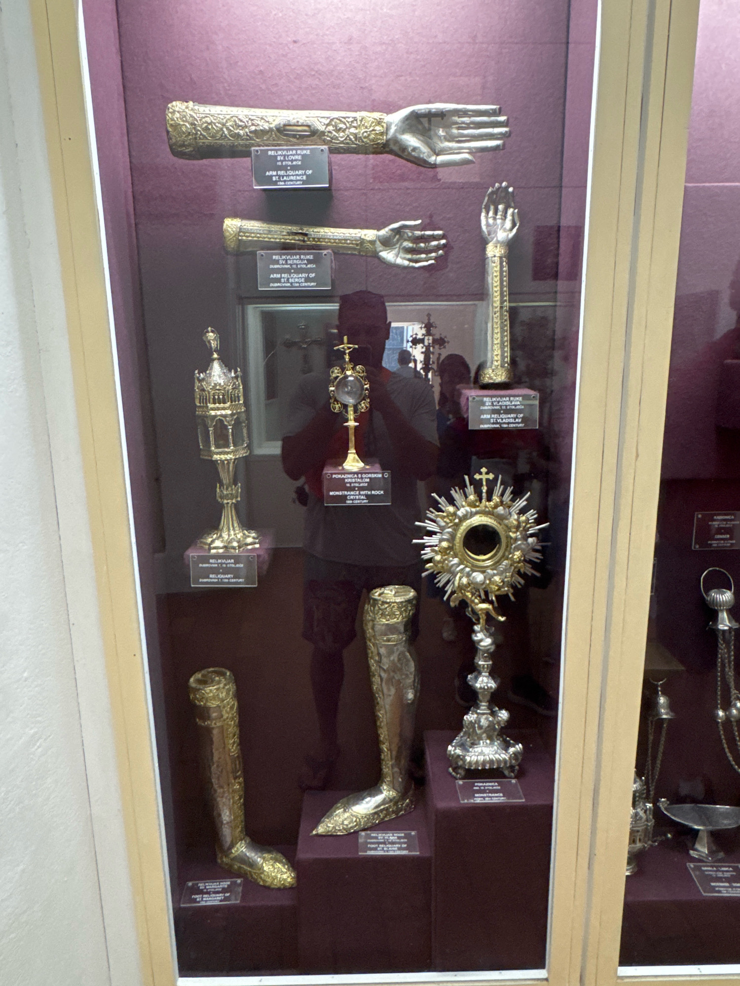This image depicts a museum display case containing religious relics in Dubrovnik, Croatia. The display case is made of glass and wood and is lit from within. The relics are primarily made of gold and silver and are ornately decorated. They include several reliquaries in the shapes of hands, feet, and a monstrance. A reliquary of Saint Lawrence, a reliquary of Saint Serge, and a reliquary of Saint Vladislav are identified by labels. A monstrance with rock crystal is also displayed. A person's reflection is visible in the glass of the display case, suggesting the photo was taken inside a museum or church. The background is a deep purple fabric. The overall scene is one of historical and religious significance, showcasing artifacts of cultural importance.
