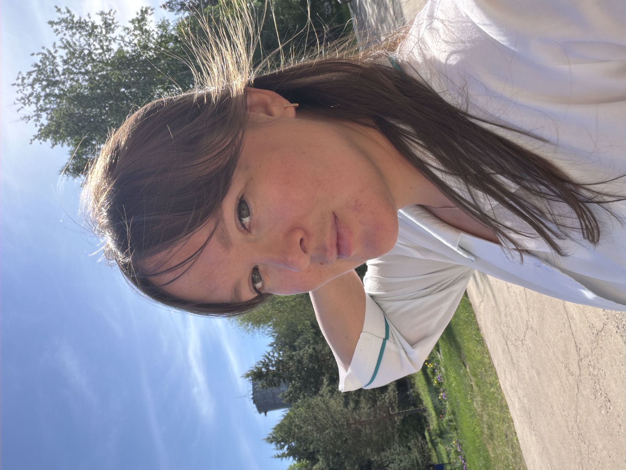 A young woman is the subject of the image, likely a selfie, as the perspective suggests she is holding the camera. She has fair skin, dark brown hair with blonde highlights, and is wearing a light-colored button-down shirt. The background reveals an outdoor setting with trees, a building, a blue sky, and a path.
Based on available information, it is presumed that the location may be in the city of Opary, Russia.