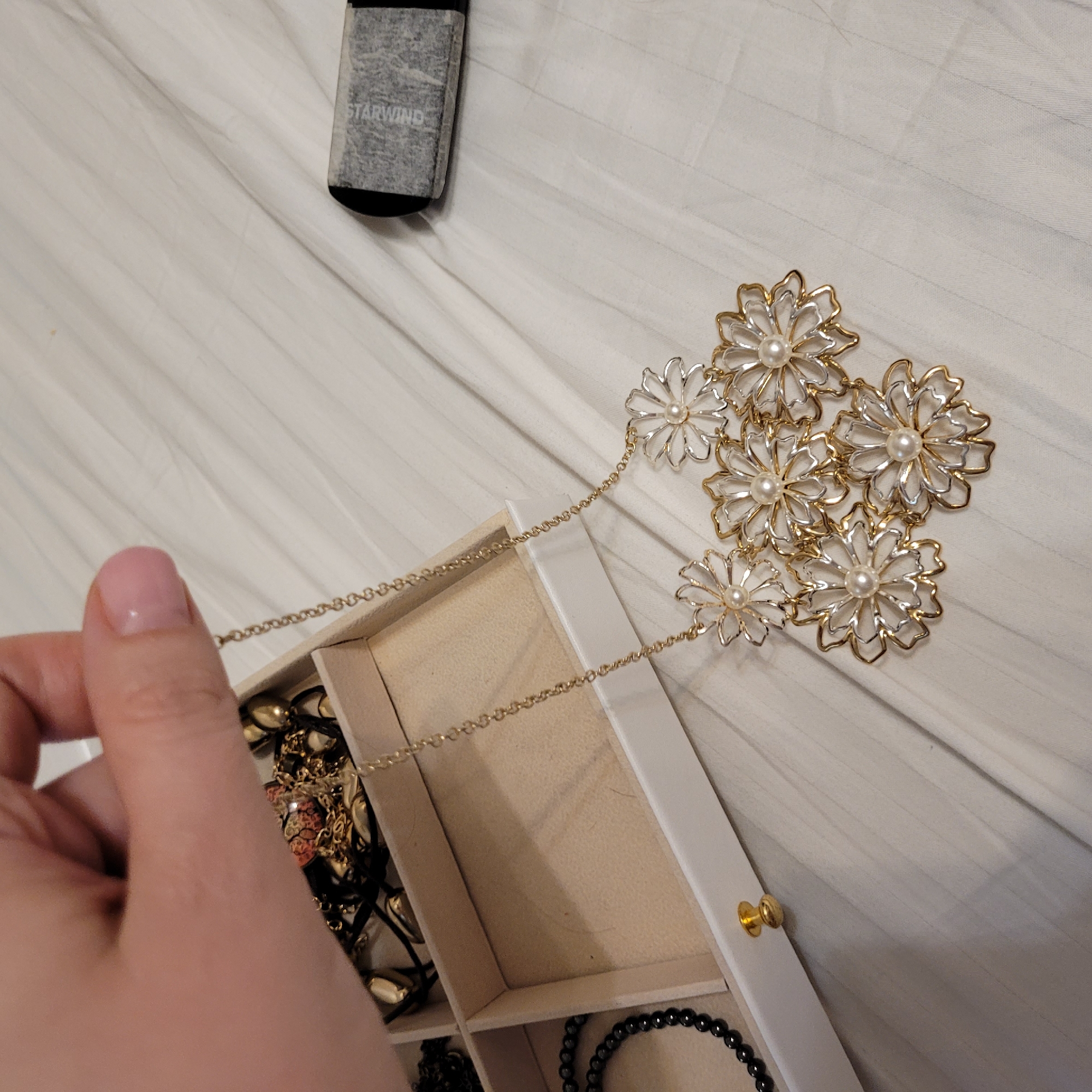An overhead shot captures a hand holding a gold-toned necklace adorned with five flower-shaped pendants, each featuring a two-toned (gold and silver-toned) outline and a central pearl. The necklace is draped over an open, white jewelry box with light-colored interior compartments. Inside the box, various other jewelry pieces are visible, including black beaded bracelets or necklaces and additional gold-toned items. The hand, with neatly manicured nails, appears to be either removing the necklace from or placing it into the box.
In the upper left corner of the frame, a black rectangular object, possibly an electronic device, is partially visible. It is wrapped in a semi-transparent, textured material, and the word "STARWIND" is legible on its surface. All these items rest on a white, horizontally-striped or pleated fabric surface, resembling bed linen or a duvet cover, suggesting an indoor setting like a bedroom. The lighting appears bright and even, indicating either daytime or well-lit indoor conditions. No specific emotions are conveyed.