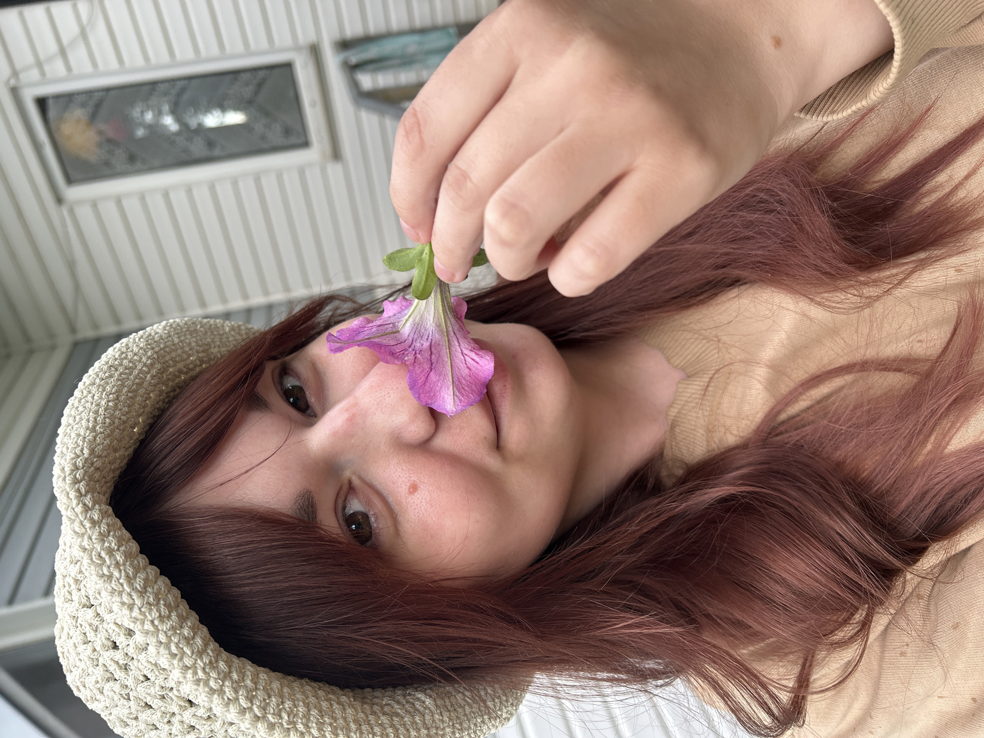 An image, rotated 90 degrees clockwise, captures a young person with fair skin, brown eyes, and long, reddish-brown hair. They are wearing a light beige knit or crocheted hat and a matching beige top. The person is holding a vibrant purple, petunia-like flower by its green stem, gently touching it near their nose or mouth area, as if smelling it. They look directly towards the viewer with a neutral to soft expression.
Notable details include a small mole on the person's cheek. The texture of the knit hat and the person's long, wavy hair are clearly visible.
The background features white vertical paneling, possibly siding or a ceiling, suggesting an indoor or covered outdoor setting. A rectangular window or skylight is visible in the upper portion of the background, showing indistinct reflections. The lighting indicates it is daytime. There is no visible text in the image. While the location context is given as Фоки, Russia, no specific visual cues within the image directly support this.