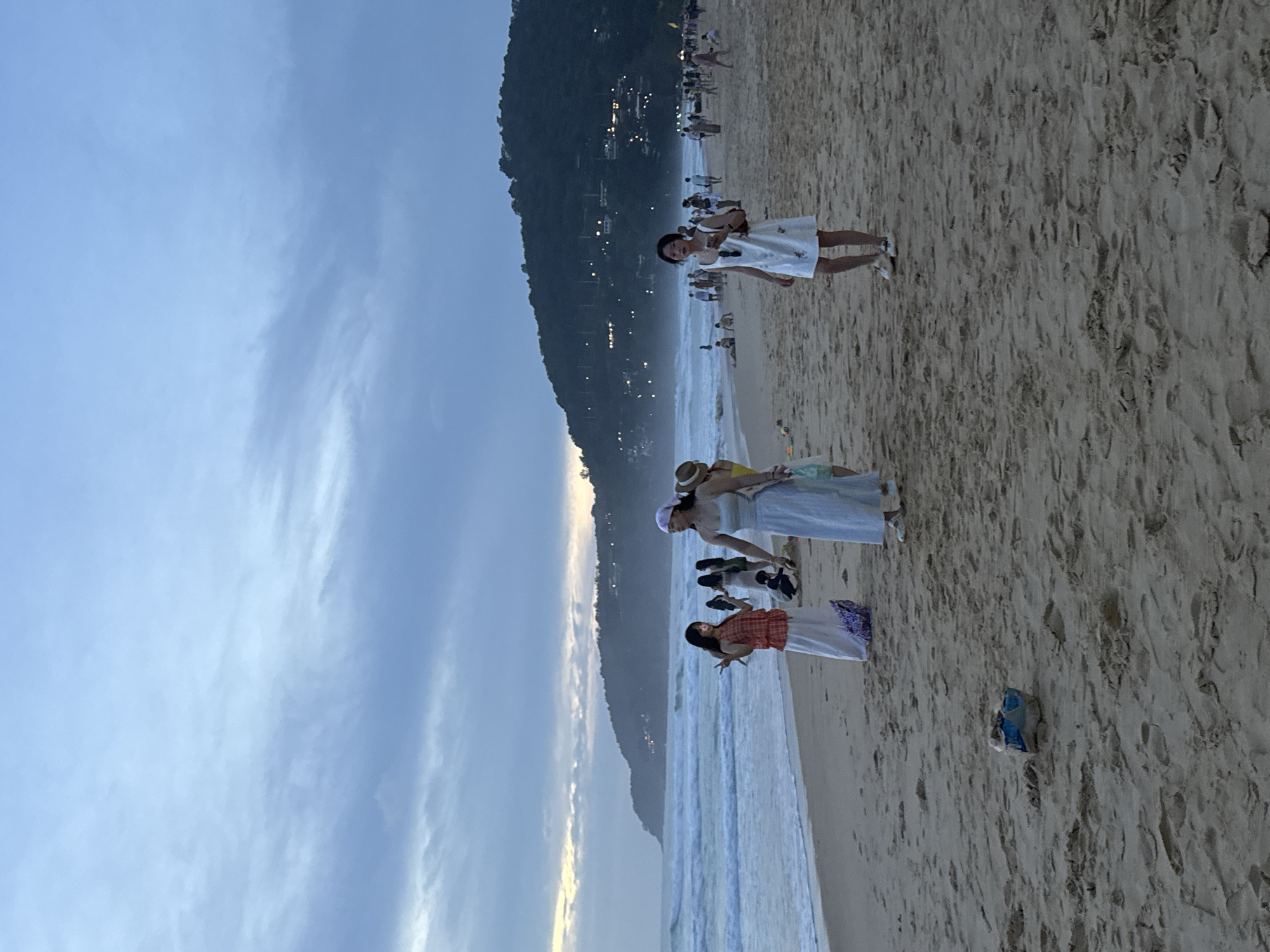 A group of people are on a sandy beach in Karon, Thailand, near dusk. The sky is overcast with some lighter areas near the horizon, suggesting sunset. Waves are breaking on the shore. In the foreground, there is a discarded blue and white object on the sand. Several individuals are walking along the beach, and some are in the water. A prominent green hill with lights on it overlooks the coastline in the background. Three women in the mid-ground are facing the camera, each wearing a dress. One woman on the far right is wearing a white dress and has a purse. The woman in the center is wearing a light blue dress and a light purple headscarf. The woman on the left is wearing a patterned orange top and a long white skirt. Several other people are visible further down the beach and in the water.