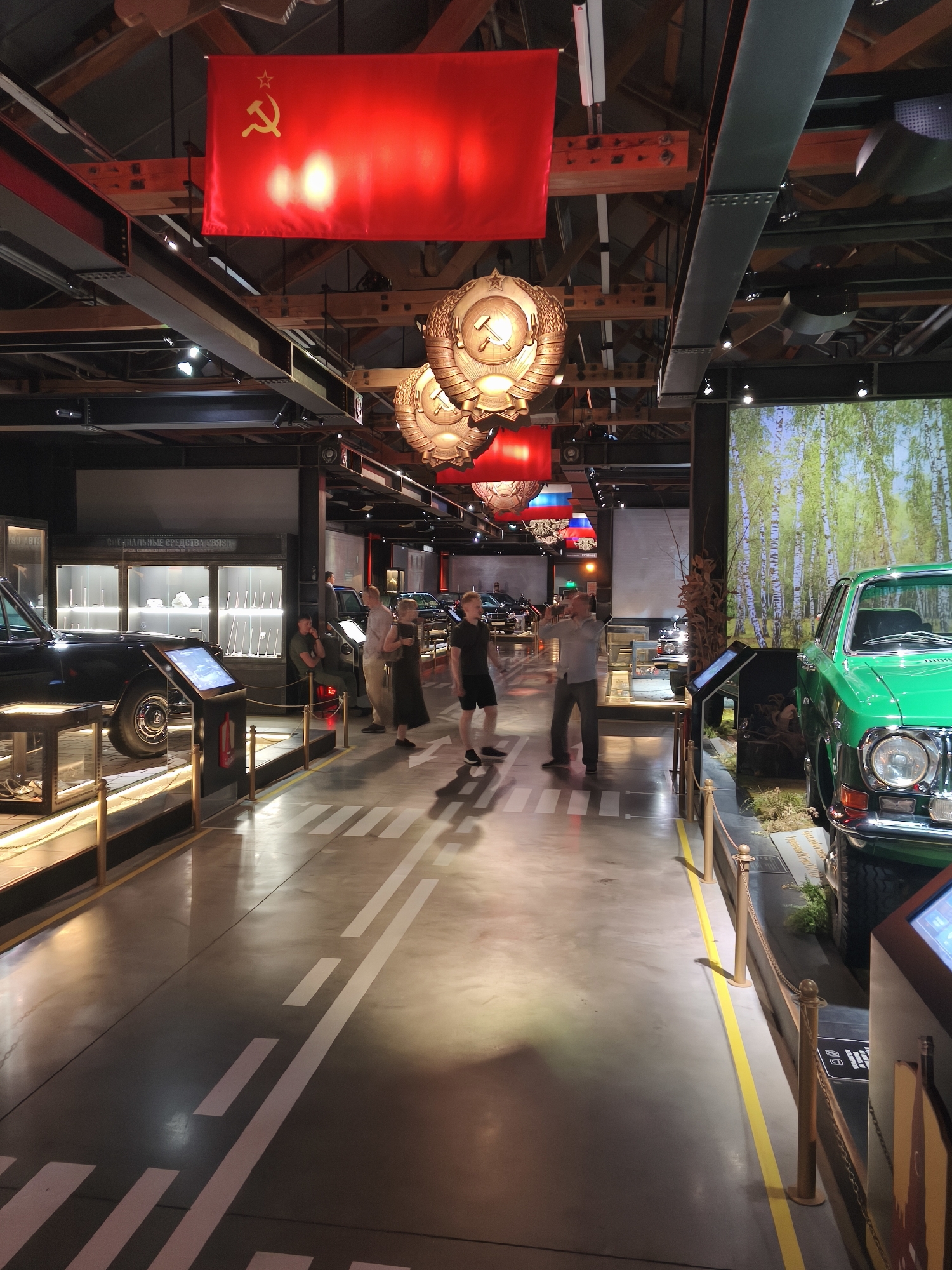 The image captures an interior exhibit, likely a museum, with a distinct Soviet theme. Prominently displayed overhead are large, red Soviet flags with the hammer and sickle symbol. Large, ornate Soviet emblems, also featuring the hammer and sickle, hang from the ceiling.
The exhibit floor features a gray concrete surface with white painted road markings. Several vintage cars are on display. To the left, a black car is partially visible, and to the right, a vibrant green car is prominently featured.
Several individuals are present in the scene, appearing to be visitors or museum staff. They are standing and observing the exhibits. One group stands near the black car, while another group is positioned further down the exhibit path, near the green car.
In the background, display cases contain various items, suggesting further exhibits or historical artifacts. The lighting is a mix of overhead industrial lighting and spotlights directed at the exhibits. A large backdrop depicting a birch tree forest is visible behind the green car, creating a diorama effect. The overall atmosphere suggests a historical exhibition, possibly related to Soviet-era automobiles or history. The context of Moscow, Russia is implied by the strong Soviet symbolism.