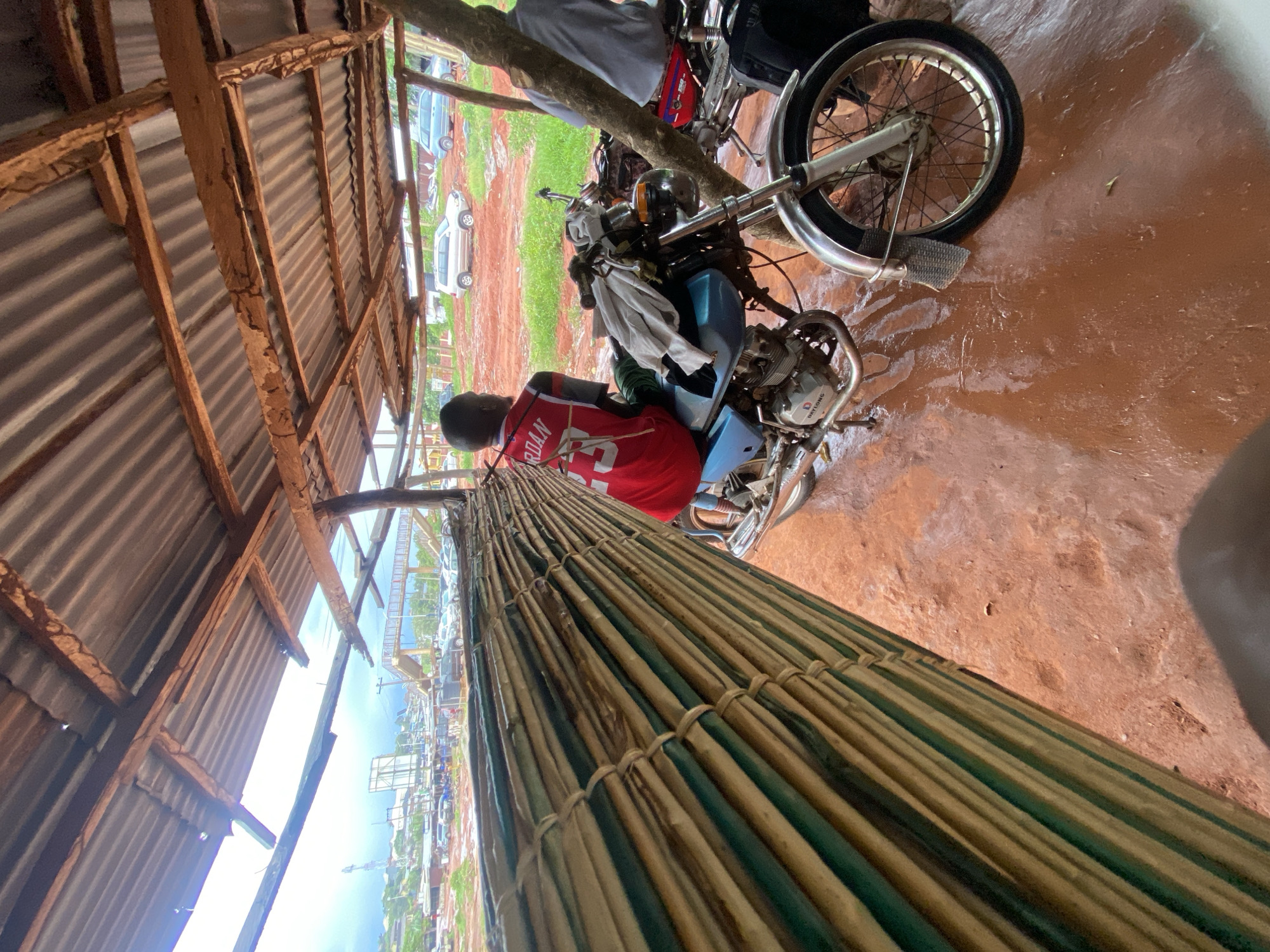 The image captures an outdoor scene, likely a temporary shelter or stall, with a person and a motorcycle as central subjects. The perspective is angled downwards, looking from above and slightly to the side.
A person, seen from the back, is seated on a light blue motorcycle. They are wearing a red jersey with the name "JORDAN" and the number "23" clearly visible on the back. The motorcycle has chrome accents and the word "DAYLONG" is visible on its engine casing. A white cloth is draped over the handlebars.
The ground beneath the motorcycle and within the shelter is a reddish-brown color, appearing somewhat wet or damp. The shelter itself has a corrugated metal roof supported by wooden beams, some of which show signs of wear. In the foreground, a large, cylindrical bundle, seemingly made of tightly woven bamboo or similar natural materials, stretches diagonally across the lower part of the frame.
Beyond the immediate shelter, the background reveals an open, earthy area with patches of green vegetation. Several cars, predominantly white and light-colored, are parked further away. In the distance, a general view of what appears to be a town or settlement is visible under a bright, overcast sky, suggesting it is daytime. A tall structure, possibly an antenna or tower, can be seen on the horizon. The overall scene suggests a functional, possibly commercial or roadside, environment in Auchi, Nigeria.
