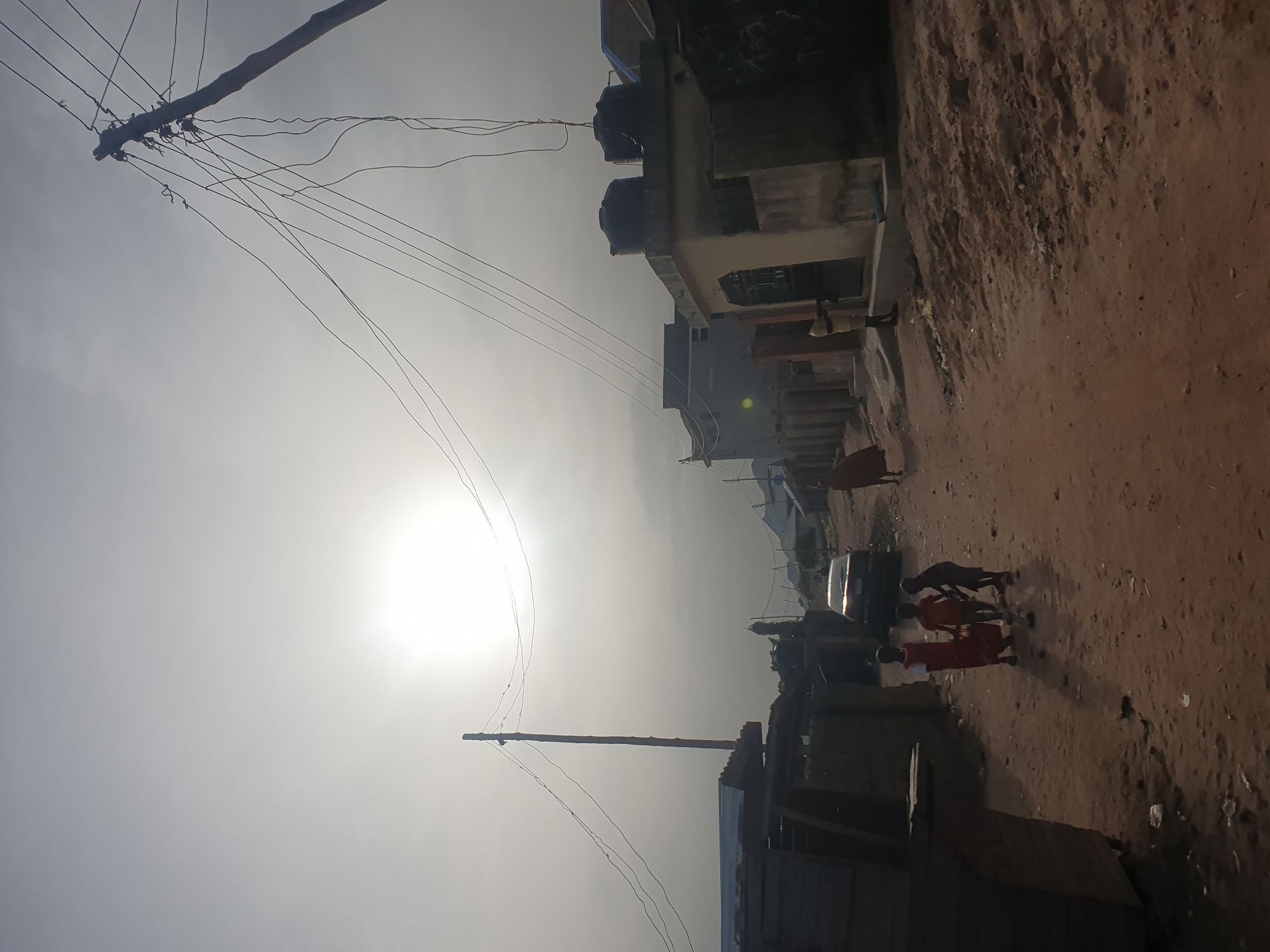 The image depicts a sun-drenched, semi-urban street scene in Lokoja, Nigeria, looking eastwards into the bright sun, which causes strong backlighting and a hazy appearance. The photo is rotated 90 degrees clockwise, showing the sky on the left and the ground on the right.
In the upper left, a utility pole stands prominently against the bright sky, featuring a dense cluster of overhead wires extending across the frame, indicating an informal or older electrical grid. Below the pole, on the right side of the frame (bottom right if correctly oriented), is a dirt road or path.
Several figures are visible on this road. In the middle-ground, a group of children, one in a bright red garment, appears to be walking or playing. One child in a red dress is seen alongside a dark, dog-like animal. Another person in a yellow top is visible further back, near a building. A dark-colored car is parked on the left side of the dirt road, partially obscured.
On the right side of the image (top if correctly oriented), a row of buildings with varied architecture lines the street. Some buildings have black water tanks on their roofs. These structures appear to be residential or commercial, with simple facades. Closer to the foreground, on the right, are smaller, possibly makeshift, wooden structures or shacks.
The scene is bathed in bright daylight, suggesting midday or early afternoon, with the sun low enough to cast long shadows and create a strong glare. The ground is unpaved, consisting of reddish-brown dirt, characteristic of many unpaved roads. No discernible text is visible on any of the structures or objects.