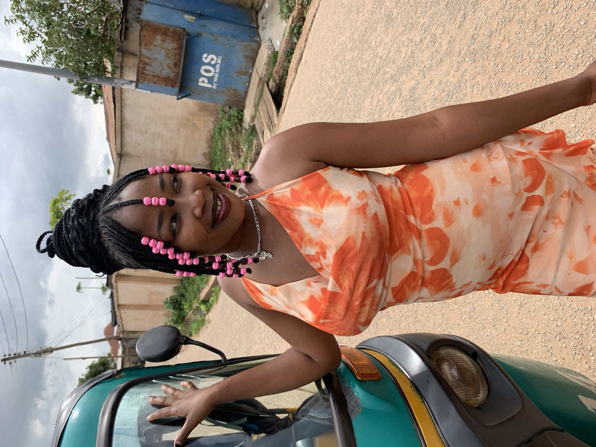 A young woman with dark skin and black braided hair adorned with pink beads, styled in an updo and framing her face, stands on an unpaved street in Jos, Nigeria. She wears a vibrant orange and cream tie-dye halter-neck dress and a silver necklace with a butterfly pendant. Her left hand rests on the windshield frame of a green three-wheeled auto-rickshaw (keke Napep), which features a black side mirror, a round headlight, and yellow trim. She is smiling and looking slightly towards the camera.
The environment under an overcast sky consists of a light brown, gravelly road. In the mid-ground, a heavily rusted blue metal bin, likely for utilities or refuse, displays the white text "POS" and "PAY YOUR NEPA BILL," indicative of Nigerian infrastructure. Adjacent to the bin, a small wooden bridge crosses a shallow ditch. Behind the woman, a beige concrete wall and a building with a rusty corrugated metal roof are visible. A wooden utility pole with overhead power lines and sparse green foliage and trees complete the background elements.