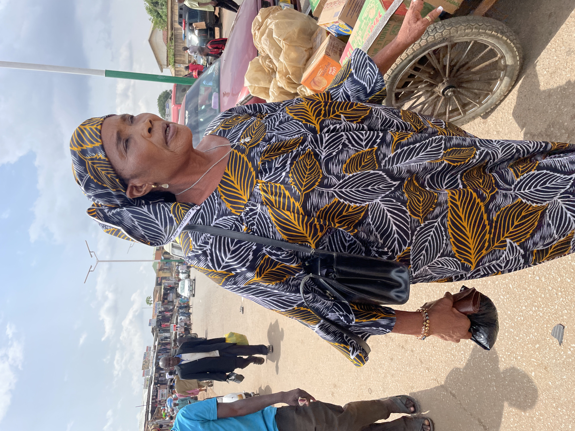 In Jos, Nigeria, an elderly woman in a black, white, and yellow leaf-patterned dress and matching headwrap stands next to a cart loaded with goods, including boxes and a large package. She is holding a black bag in her right hand, a black purse hangs from her shoulder, and she wears a bracelet and necklace. She gazes upward. The cart has a spoked wheel. Behind her, a red car is partially visible, along with other people and vehicles along the roadside. A man in a suit walks by. The sky is partially cloudy. The environment appears to be a busy street with shops and vendors.