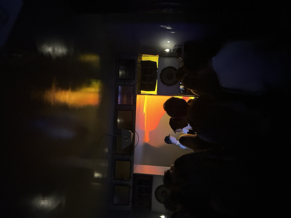 A dimly lit room features a row of square lighting fixtures on a wall, reflecting yellow and orange hues on the floor. A speaker system is visible near the light fixtures. Silhouetted figures are positioned near a counter, seemingly observing or interacting with the space. The dark environment obscures details of the people and the room's contents.