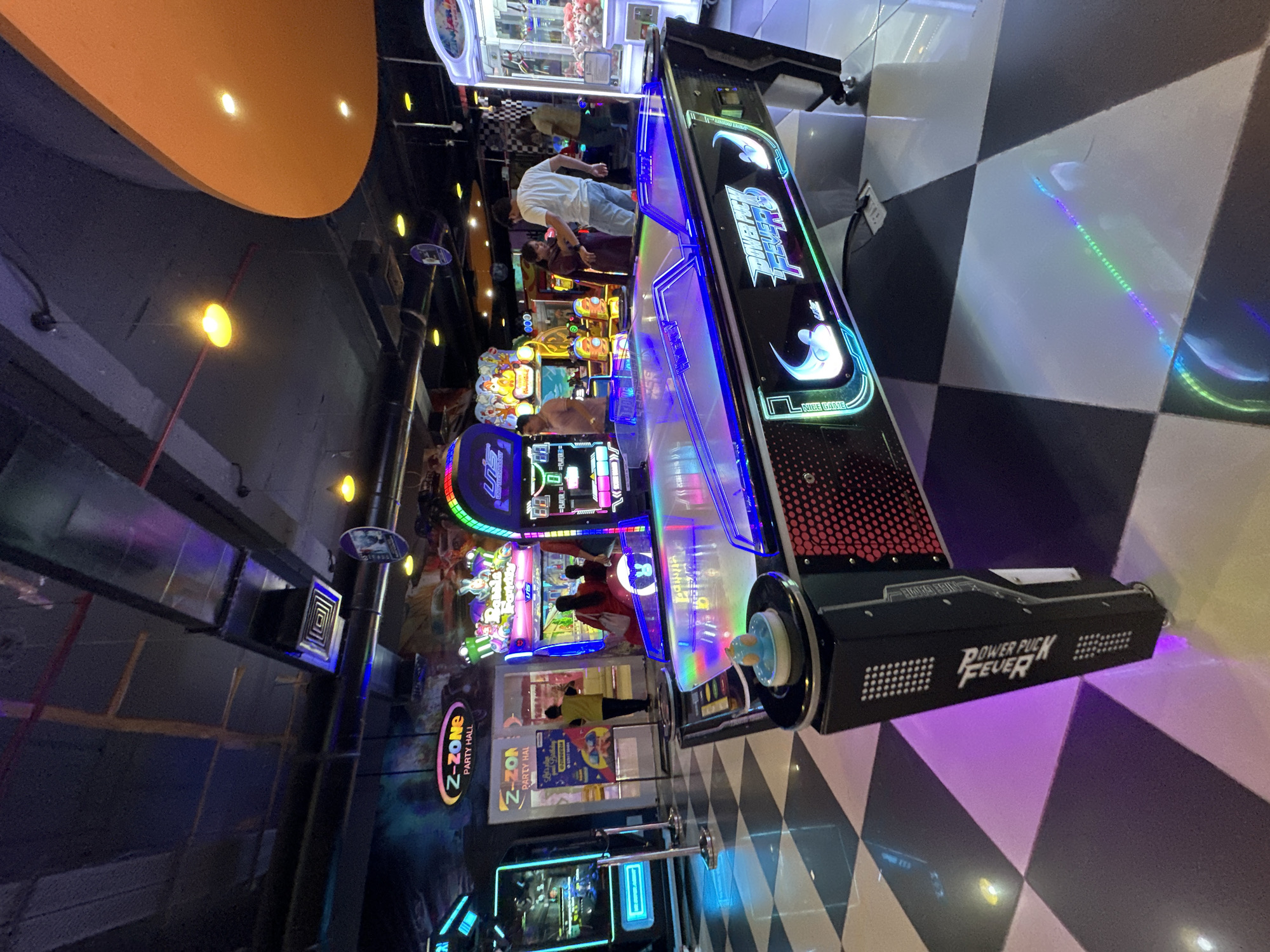 The photograph captures an interior scene in what appears to be an entertainment or arcade center, possibly located in Kalyan-Dombivli, India, though this cannot be definitively confirmed from the image alone. The primary focus is a "Power Puck Fever" arcade game, standing prominently on a black and white checkered floor. The game features illuminated elements and bold lettering. Several other arcade machines are visible in the background, suggesting a bustling entertainment environment. People are seen interacting with the games in the background. Overhead, an orange ceiling structure is apparent along with industrial ductwork. The overall lighting is a mix of ambient and directed light, creating a vibrant atmosphere.