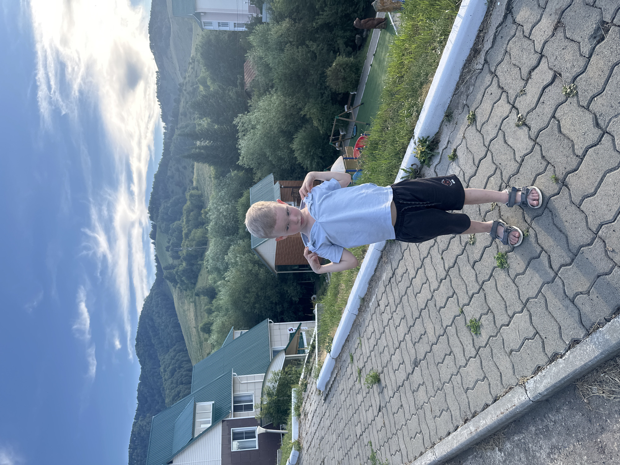 A young boy stands on a paved pathway in what appears to be a resort area. He is wearing a light blue polo shirt and dark shorts, with grey sandals on his feet. The boy is looking down and holding the collar of his shirt with both hands. The pathway is made of interlocking grey pavers and slopes gently uphill. To the left of the pathway, there are several buildings with green roofs. In the background, lush green hills and trees are visible, with a cloudy sky overhead. To the right of the pathway, there is a playground with swings and a slide, and further back, a large building. There are also some decorative bear statues visible on the right side of the image. The overall impression is a scenic, outdoor location likely in a mountainous region, consistent with the provided location information of Danilovka (Altai), Russia, known for its natural beauty and recreational facilities.