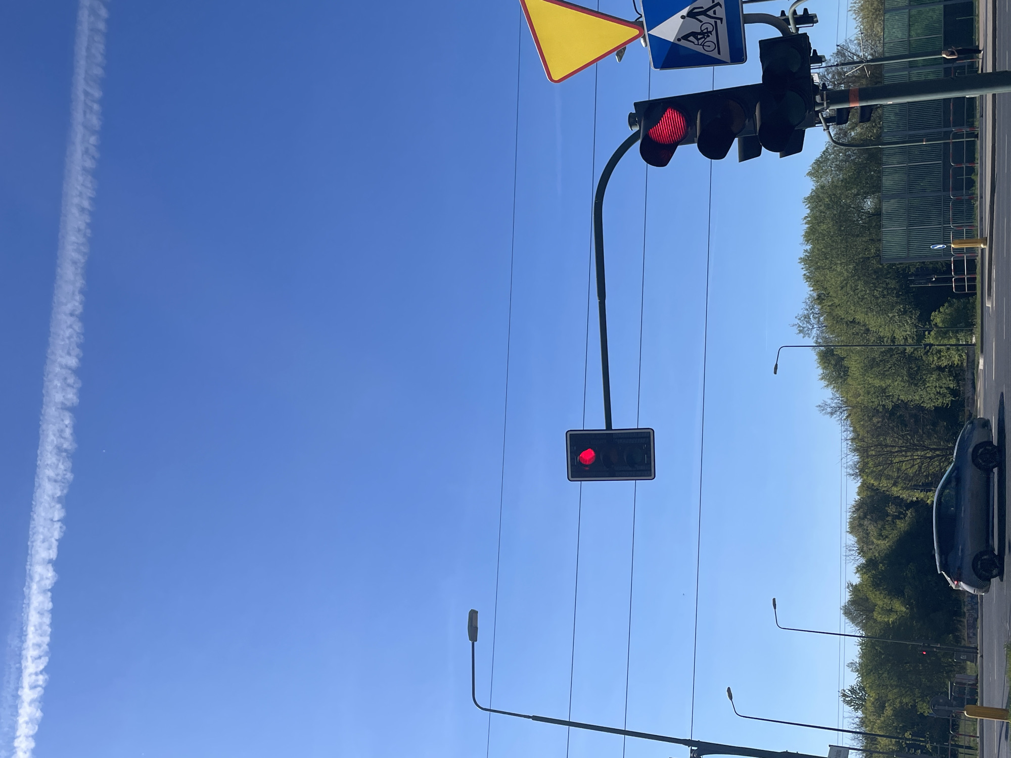 The image depicts a street scene in an unknown city. The sky is a clear blue with a contrail visible in the upper left. A traffic light is showing red in the upper right, and a second, smaller traffic light also shows red further down the frame. A road sign is located above the main traffic light. Trees line the street, and a parked car is on the right side of the image. Several streetlights extend from the left side of the frame. Overhead wires are strung across the sky.