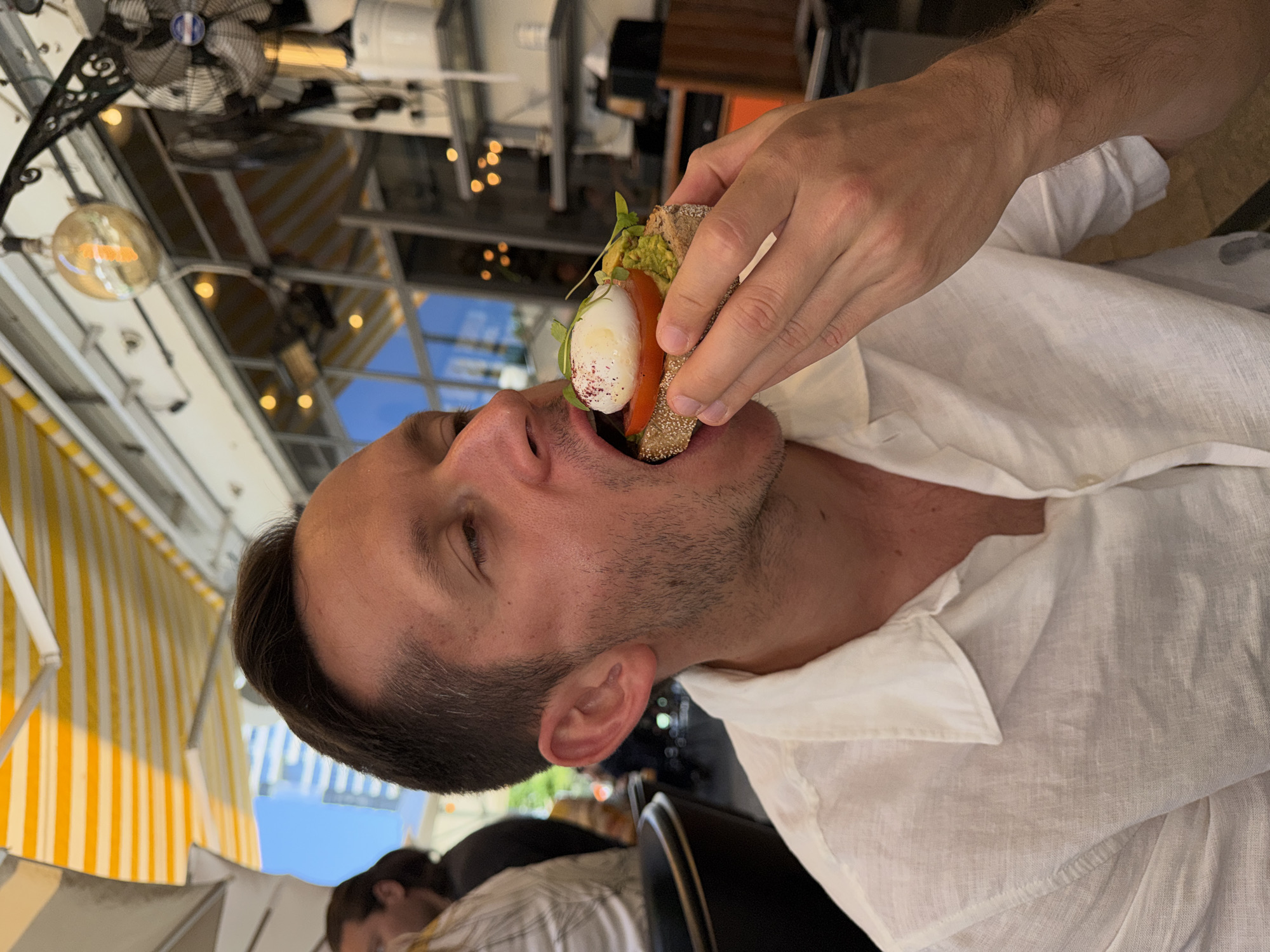 A man is eating a sandwich in what appears to be a Tel Aviv, Israel restaurant. The sandwich contains a poached egg, tomato, and avocado and is held in his right hand. He is wearing a white, button-down shirt. The background includes a yellow and white striped awning, outdoor lighting fixtures, and an indoor fan.