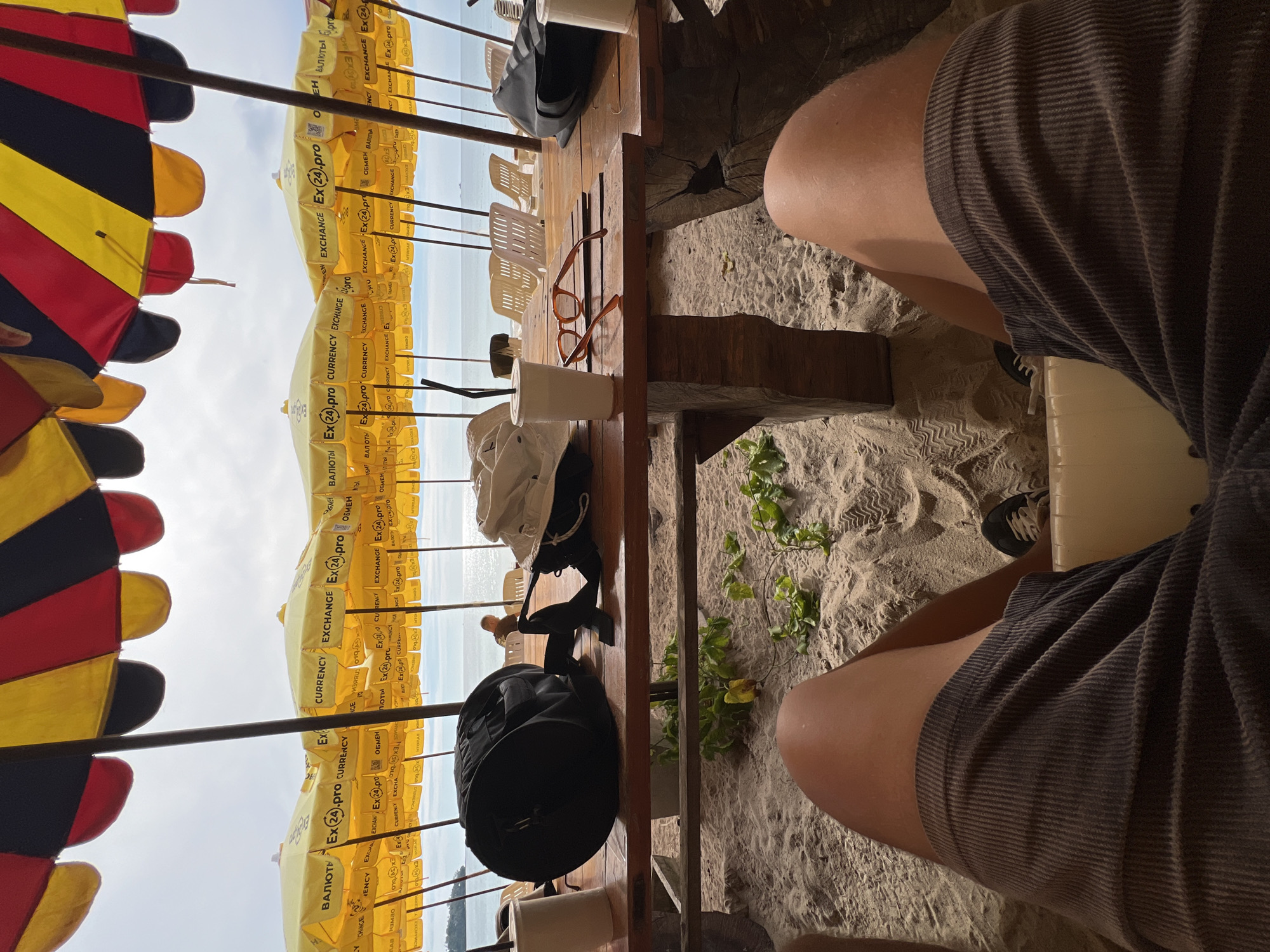 A ground-level shot looking out from under beach umbrellas onto a sandy area, possibly a beachside cafe or seating area. The umbrellas in the foreground are multi-colored (red, yellow, black) and yellow with text and are stacked close together, creating a dense canopy. Beyond the umbrellas, there is a glimpse of the sea and a cloudy sky. In the right foreground, there is a person's legs in brown shorts, with shoes visible on the sand. On a wooden table, there is a pair of glasses, a cup, a black bag, and possibly other items. The sandy ground is visible beneath the table.