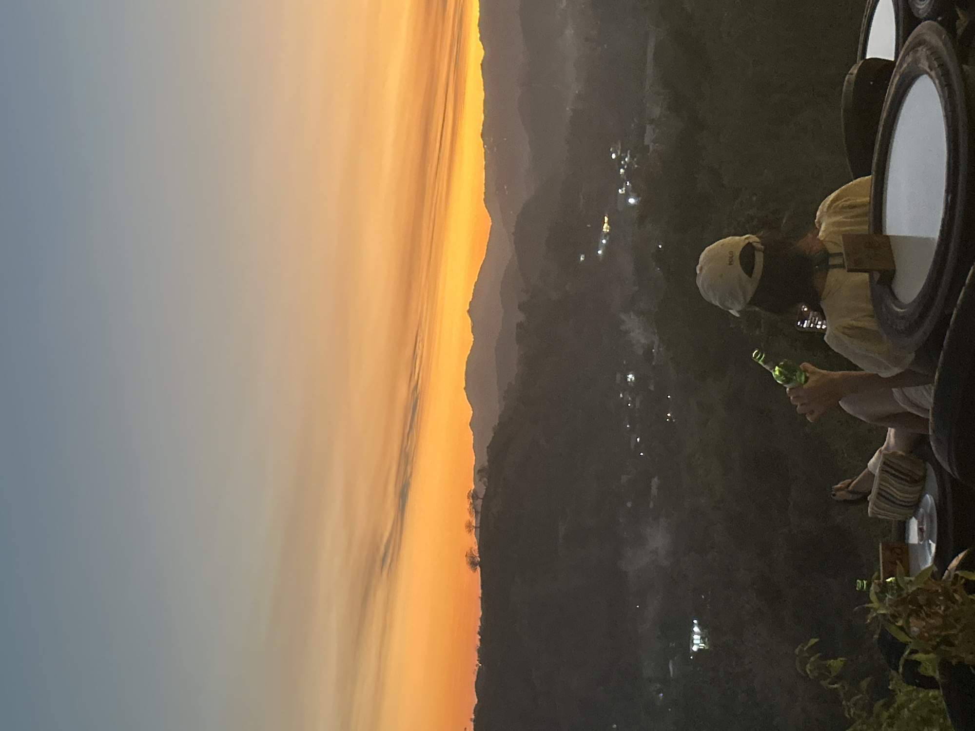 The media file depicts a person holding a green bottle, seated on what appears to be stacked tires, overlooking a mountainous landscape at dusk. The sky is painted with a vibrant gradient of orange and yellow hues, transitioning to a soft blue at the top, suggesting a sunset or sunrise. Below the horizon, the landscape is shrouded in darkness, dotted with small lights indicating a settlement or village in the valley. The person is wearing a white cap and a light-colored top, and their legs are visible, clad in striped footwear. The overall scene conveys a sense of relaxation and contemplation, with the vastness of the landscape and the fading light creating a serene atmosphere. The setting appears to be an elevated viewpoint, possibly a roadside stop or a scenic overlook in Kuta, Indonesia. No visible text is present in the media file.
