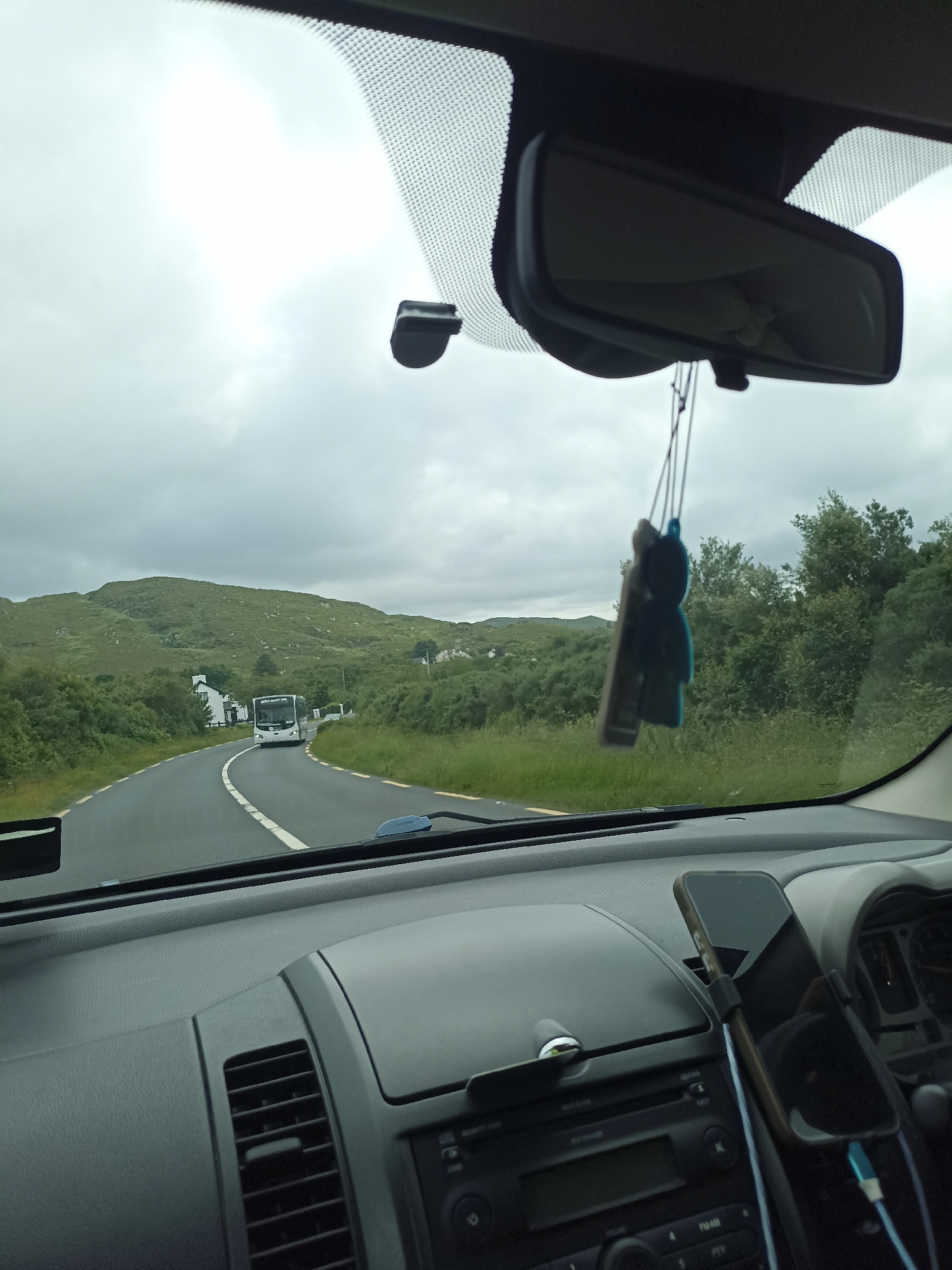 The image captures a view from inside a car driving on a winding road in Crolly, Ireland. The scene is dominated by lush green rolling hills covered in vegetation, typical of the Irish landscape. A cloudy sky suggests overcast weather.
In the distance, a white bus is approaching the car on the road. The road itself is asphalt with white and yellow lines indicating lanes. There are scattered houses and buildings visible amidst the greenery, hinting at a rural or semi-rural setting. Dense green trees line the sides of the road, creating a natural tunnel effect in some areas.
Inside the car, the dashboard is visible, featuring a car stereo and an air vent. A black car accessory, possibly an air freshener shaped like a figure, hangs from the rearview mirror. A smartphone is mounted on the dashboard, its screen reflecting the car's interior. The time of day appears to be daytime, given the ambient light, but the overcast sky contributes to a somewhat muted light. The overall impression is a scenic drive through the Irish countryside under a cloudy sky.