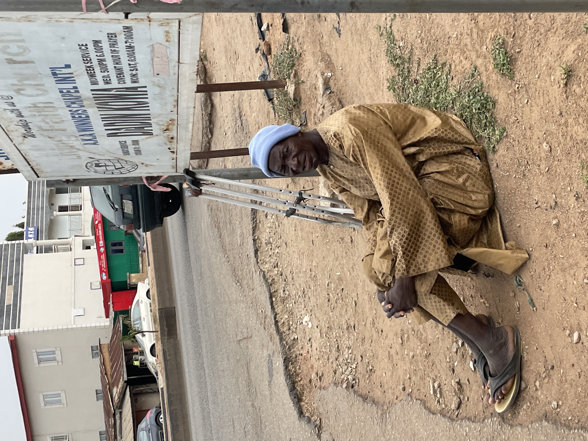 An elderly man with a blue cap is sitting on the ground beside a sign for "AKA Winners Chapel Int'l" in Jos, Nigeria. He is wearing a brown patterned outfit and black sandals, and is holding his hands together near his knees. He appears to be using crutches, which are propped up beside him and the sign. The sign advertises midweek services and covenant hour of prayer. The ground is dirt and gravel, and there are buildings and cars in the background.