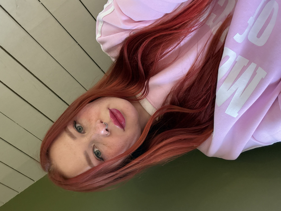 An image depicts a close-up portrait of a single female subject in an indoor setting. The subject has long, reddish-orange hair, light-colored eyes, and a fair complexion with visible freckles on her nose and cheeks. She wears a silver-colored septum piercing and bright pink lipstick. Her attire consists of a light pink long-sleeved garment, possibly a sweatshirt, featuring two parallel white stripes on the upper right shoulder/sleeve area and partially visible white text, possibly "MOSC," on the garment's front or lower sleeve. A white collar or undershirt is visible beneath the pink garment. The subject is looking directly at the camera, with her head slightly tilted. The background consists of two distinct sections: a solid, dark olive-green wall or surface on the left and a light-colored, horizontally paneled wooden wall or ceiling on the right and top. There are no discernible features within the image to identify a specific location in Rostov-on-Don, Russia; the scene appears to be a generic interior space.