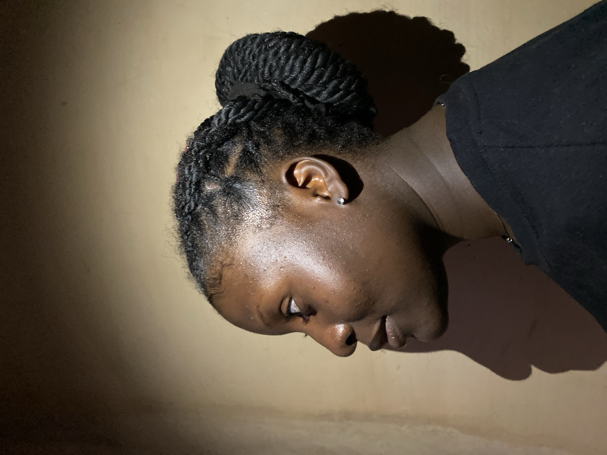A dark-skinned person with braided hair styled in a bun is depicted in a close-up, low-light photograph. The person is wearing a dark-colored garment and has a small earring in their left ear. Their head is tilted downward and to the side. The background is a plain wall, variably lit, casting a shadow behind their head. Location information regarding Kuru Dungus, Nigeria, is not discernible from the image.