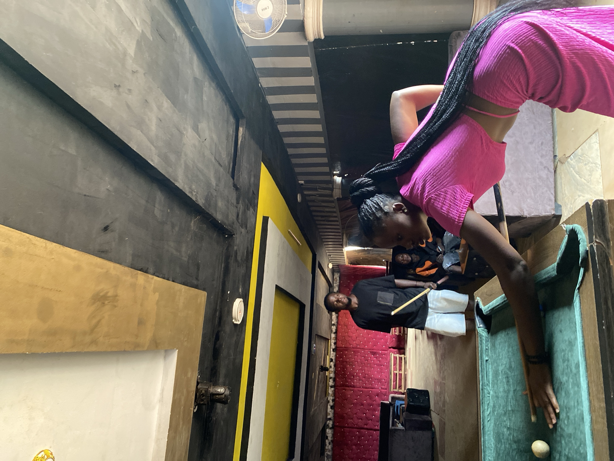 A rotated indoor shot shows a pool game in Durumi, Nigeria. A woman in a pink outfit bends over a green-felted pool table with a billiard ball on it, her arm extended, hand near the ball. Her long braided hair hangs down towards the table. Another man in a black shirt and white pants stands to the side, holding a pool cue. A third and fourth person sits on a sofa behind the man in black and white, and behind the woman in pink.
The background shows a red cushioned wall, black walls with gold and yellow accents, a ceiling fan, and a striped ceiling. The room is lit with overhead lighting. A door is visible in the background.