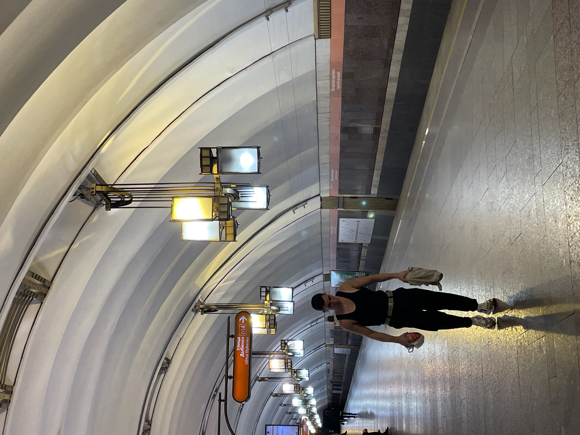 The image depicts the interior of a metro station. A person wearing a black tank top, black pants, a belt, and light-colored sneakers is standing on the platform. They are holding items in both hands. Above, an arched ceiling with multiple rows of lights dominates the scene. There is signage with text, partially readable as "Улица Дыбенко" and "Ulitsa Dybenko." The station wall features a design with contrasting dark and light panels. Other individuals are visible in the background.
